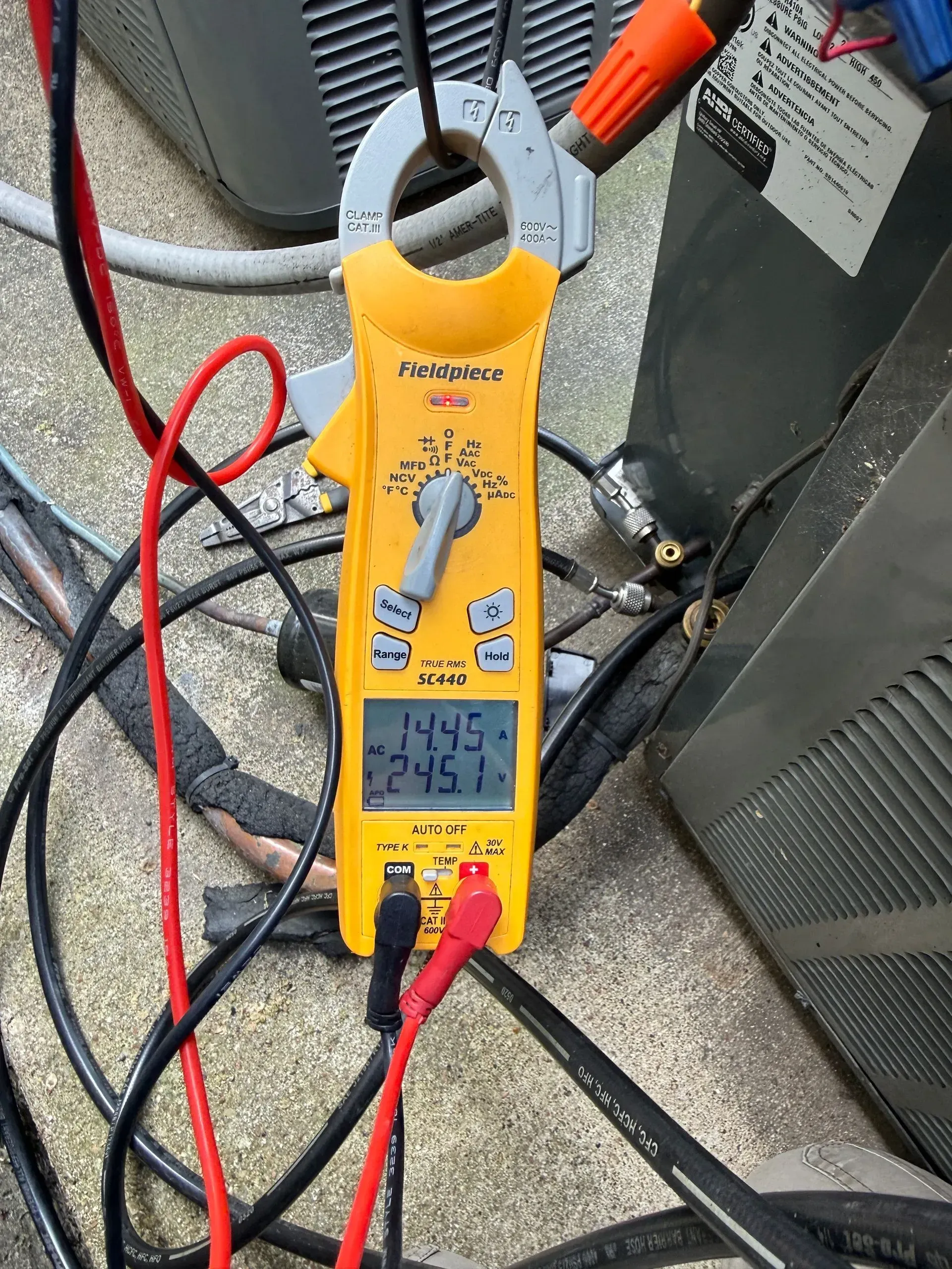 Yellow clamp multimeter on gray equipment with red and black test leads, displaying 14.5 volts.