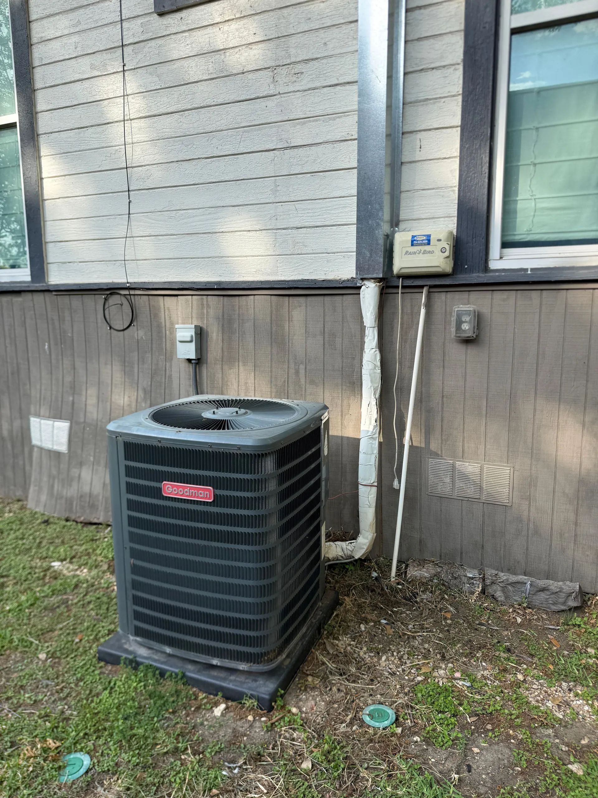 Outdoor HVAC unit beside a gray building wall with utility boxes and grassy ground