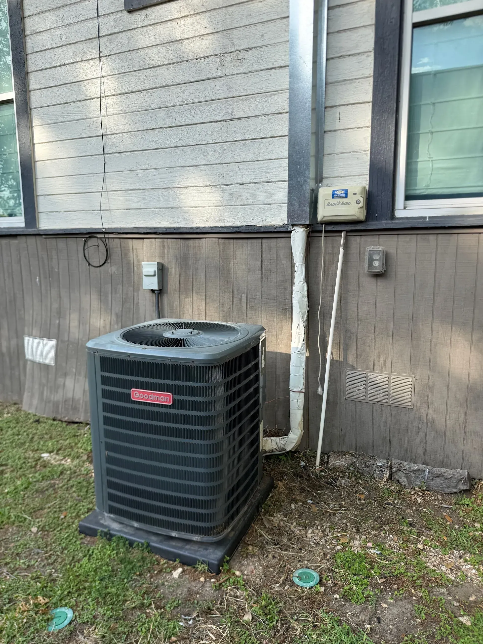 Outdoor HVAC condenser beside a house wall, with utility meters and pipes on the exterior siding.