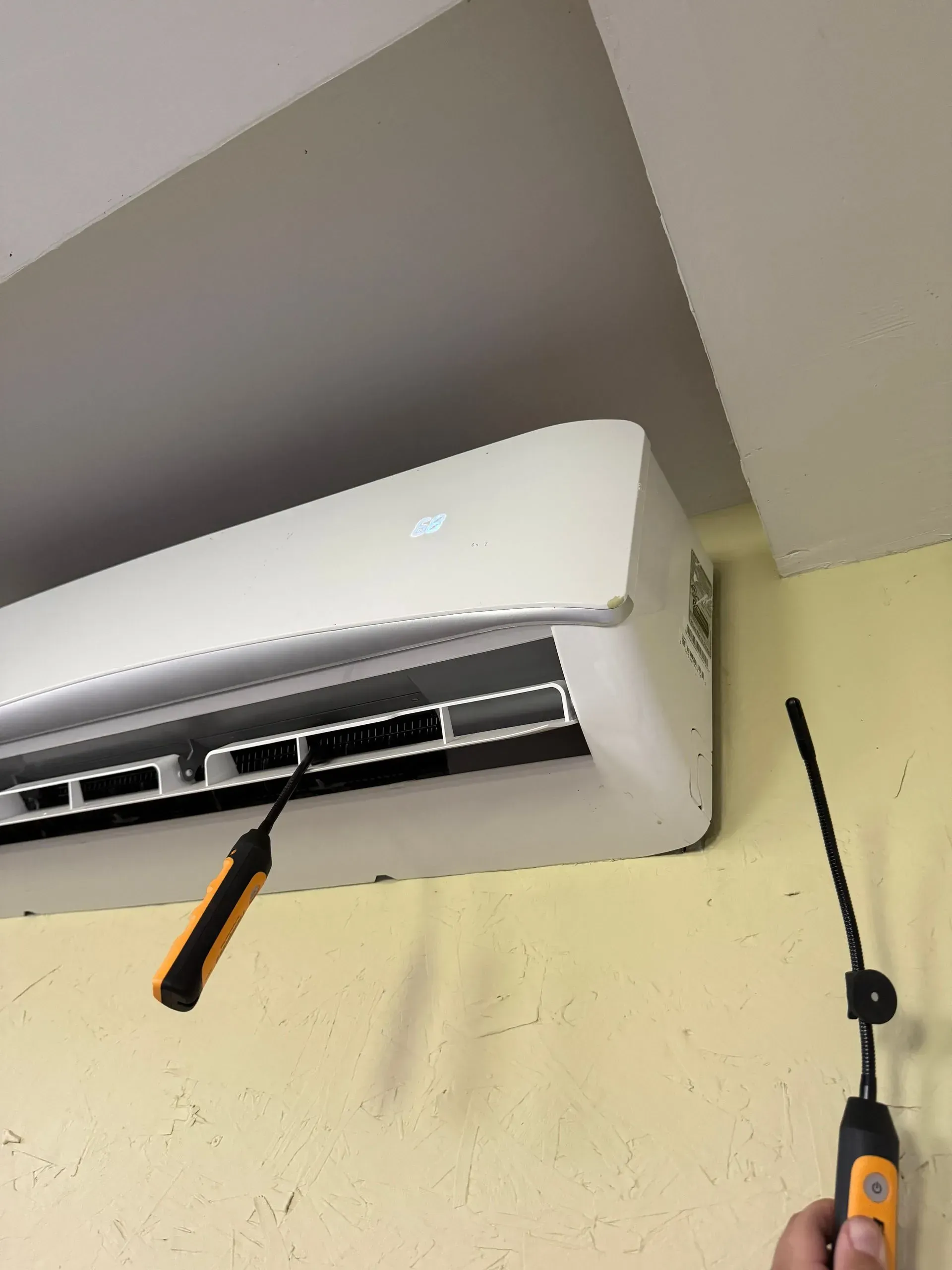 Wall-mounted white air conditioner being cleaned with a long-handled brush in a room corner.