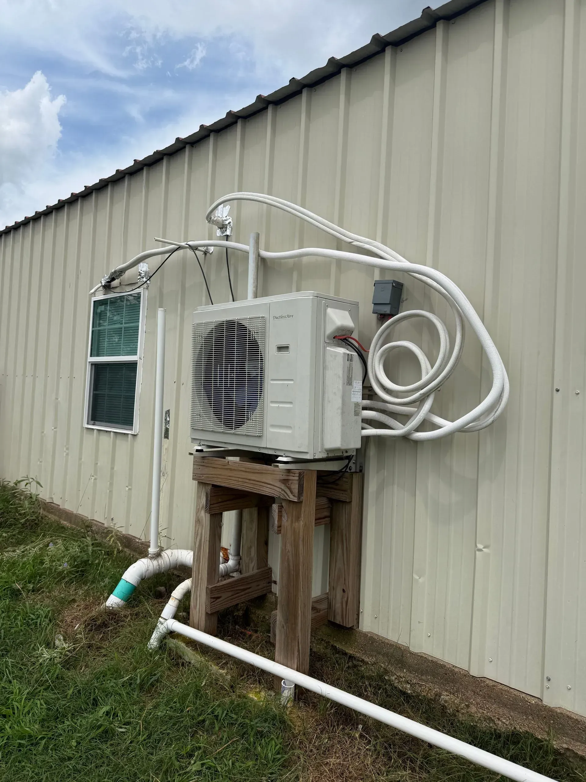 Wall-mounted air conditioner unit on a beige building with exposed white piping and grass below.
