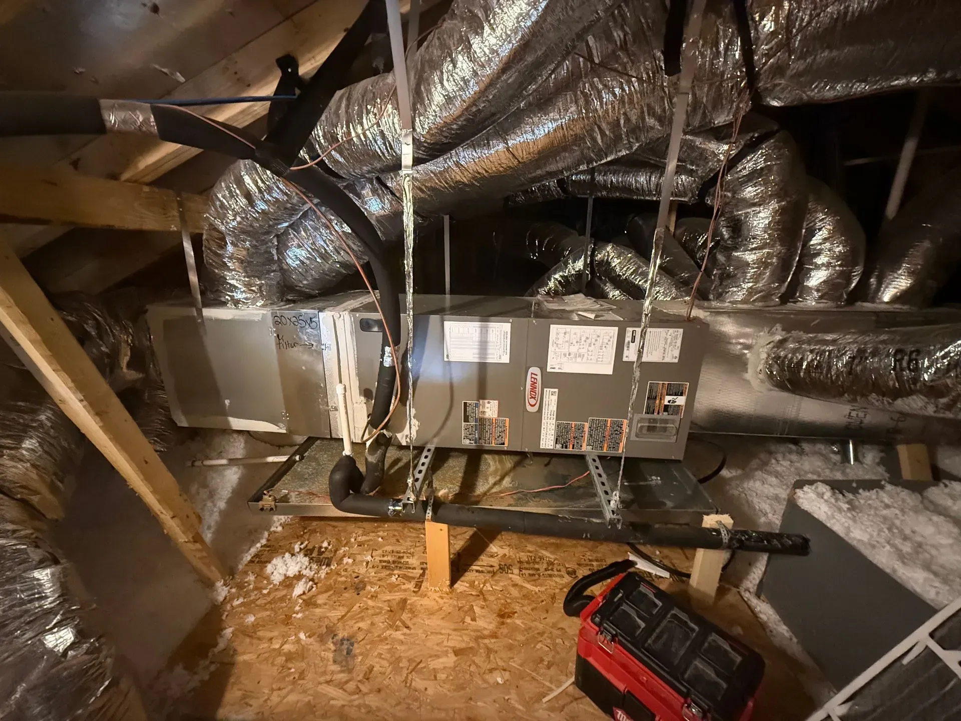 Attic HVAC unit with insulated ductwork and exposed beams; a red tool sits on the floor below.