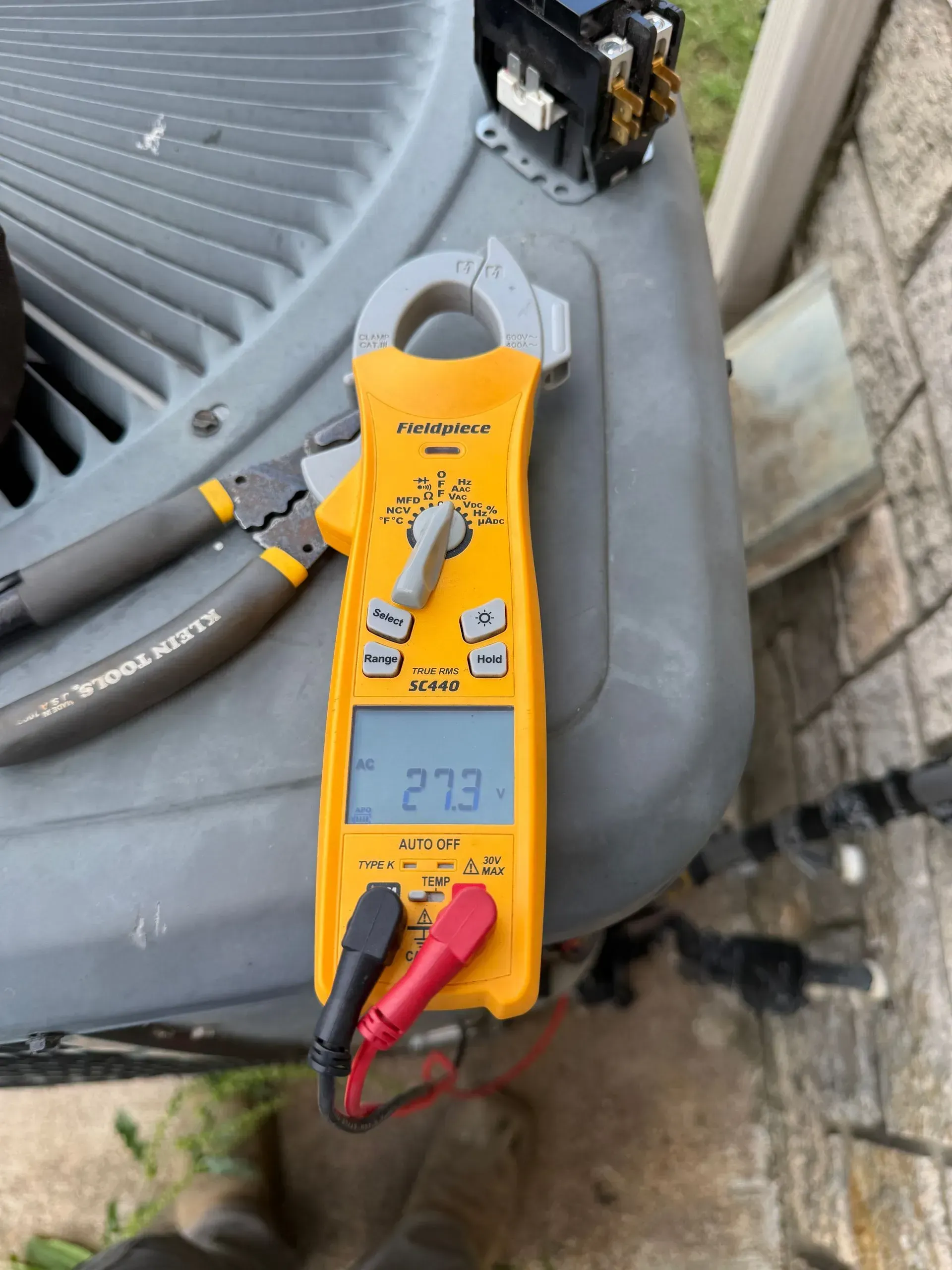 Yellow clamp meter measuring a gray HVAC unit, with red and black probes attached.