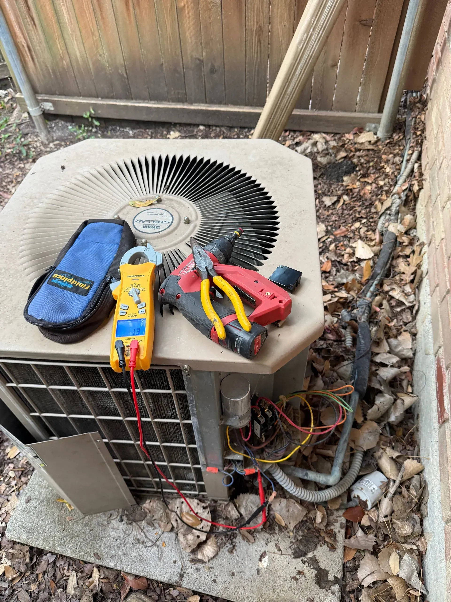 Damaged outdoor air conditioner unit with multimeter and testing tools on top, wires exposed below