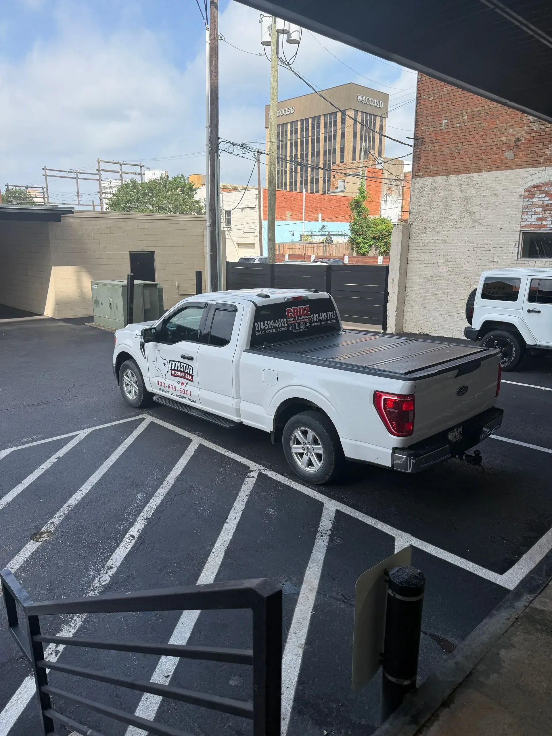 White pickup truck parked in a striped lot beside a brick building and black bollards.
