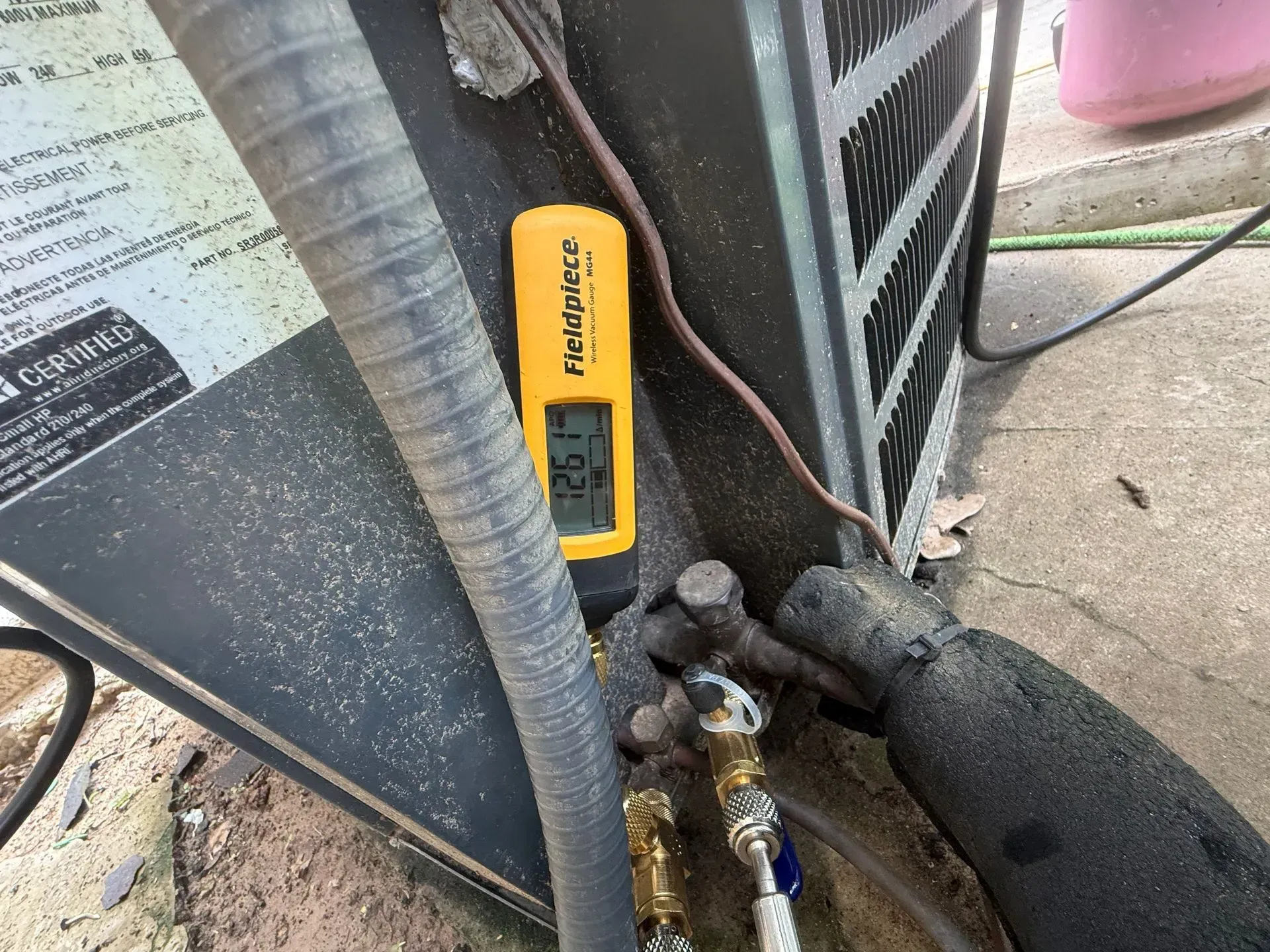 Handheld yellow digital thermometer near HVAC unit with connected hoses and a gloved hand