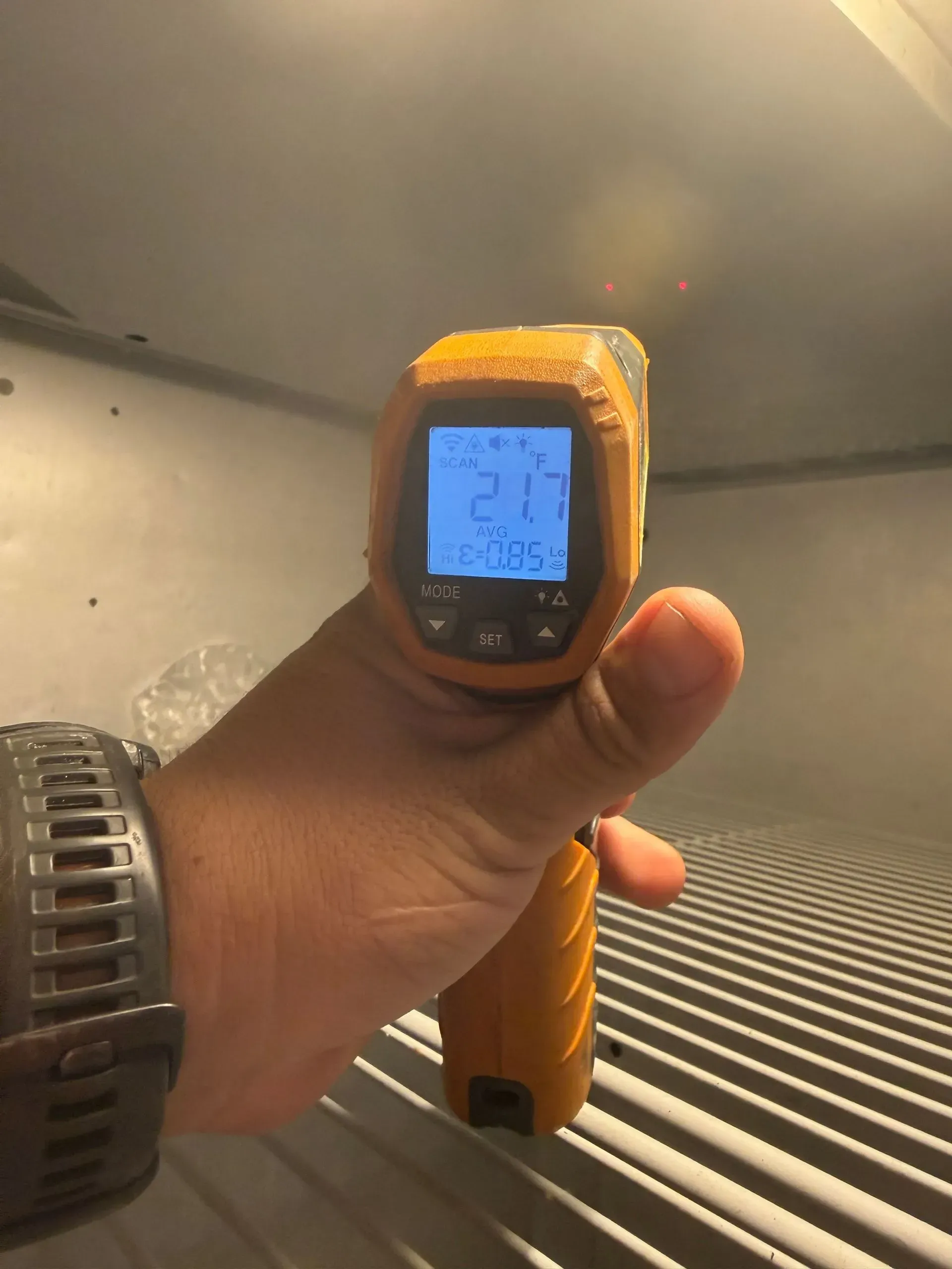 Hand holding an orange infrared thermometer aimed at a black-and-white striped surface indoors.