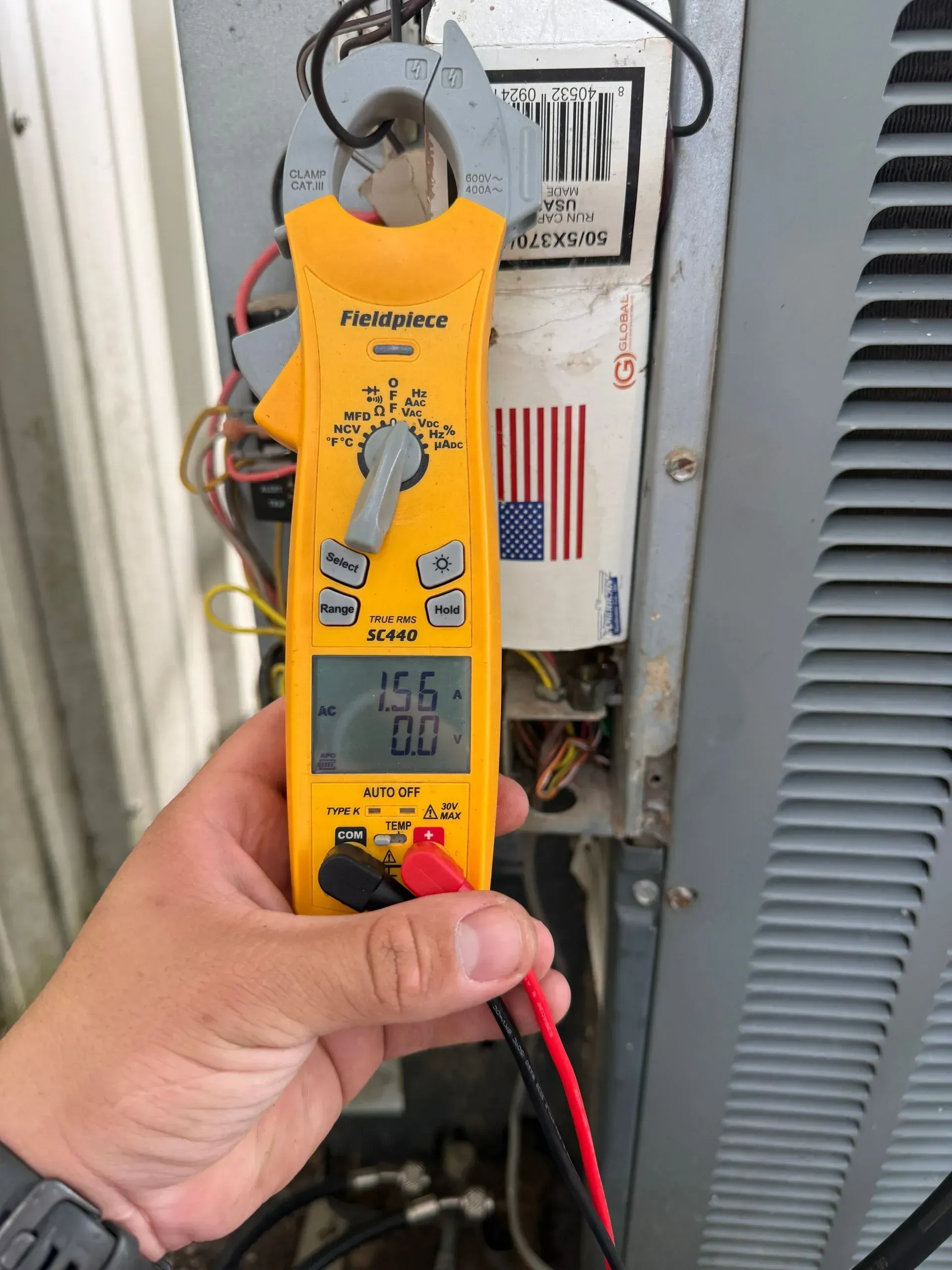 Hand holding a yellow digital clamp meter testing an electrical panel with exposed wiring.