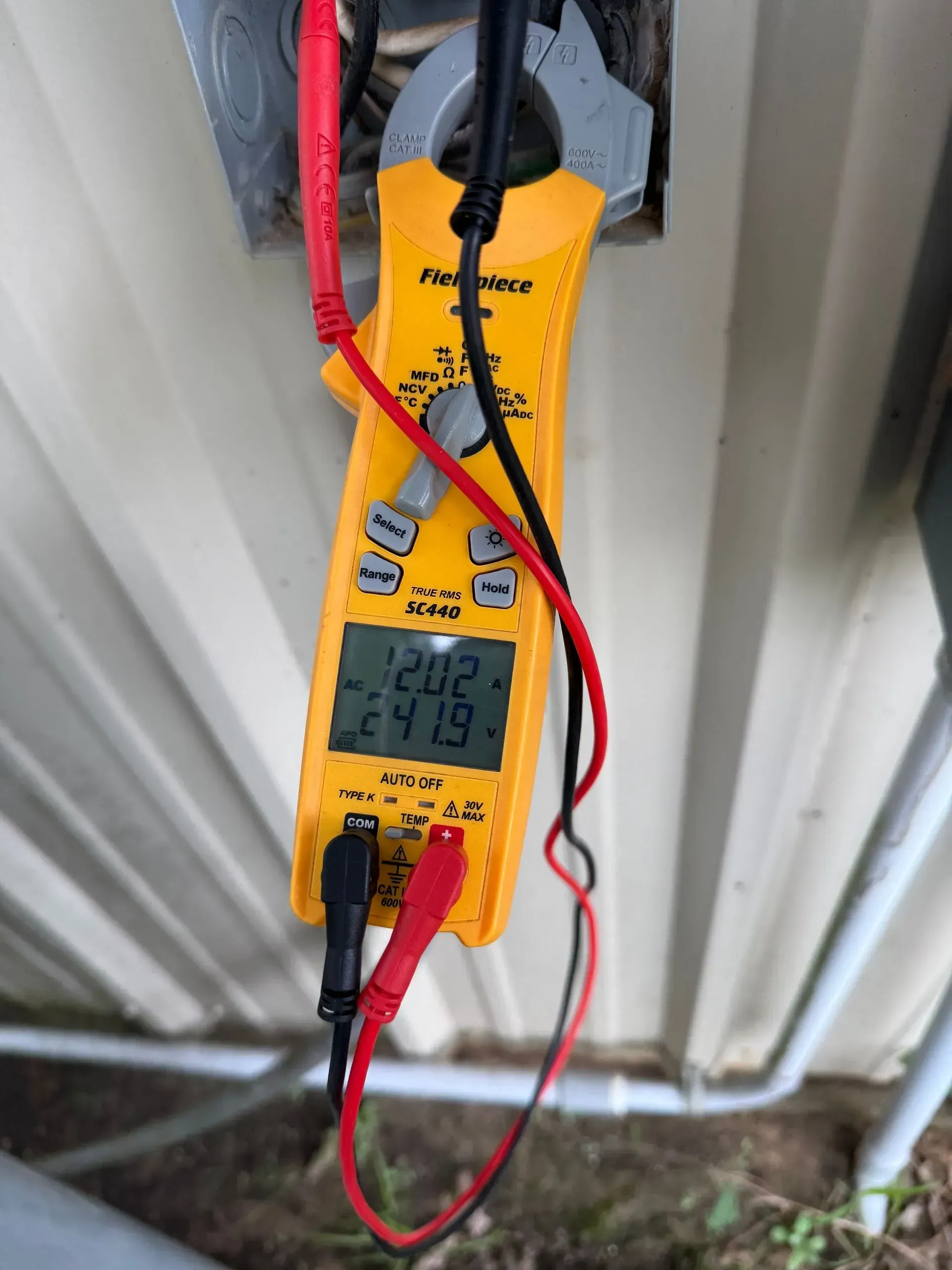 Yellow digital multimeter with red probes attached to an outdoor electrical panel