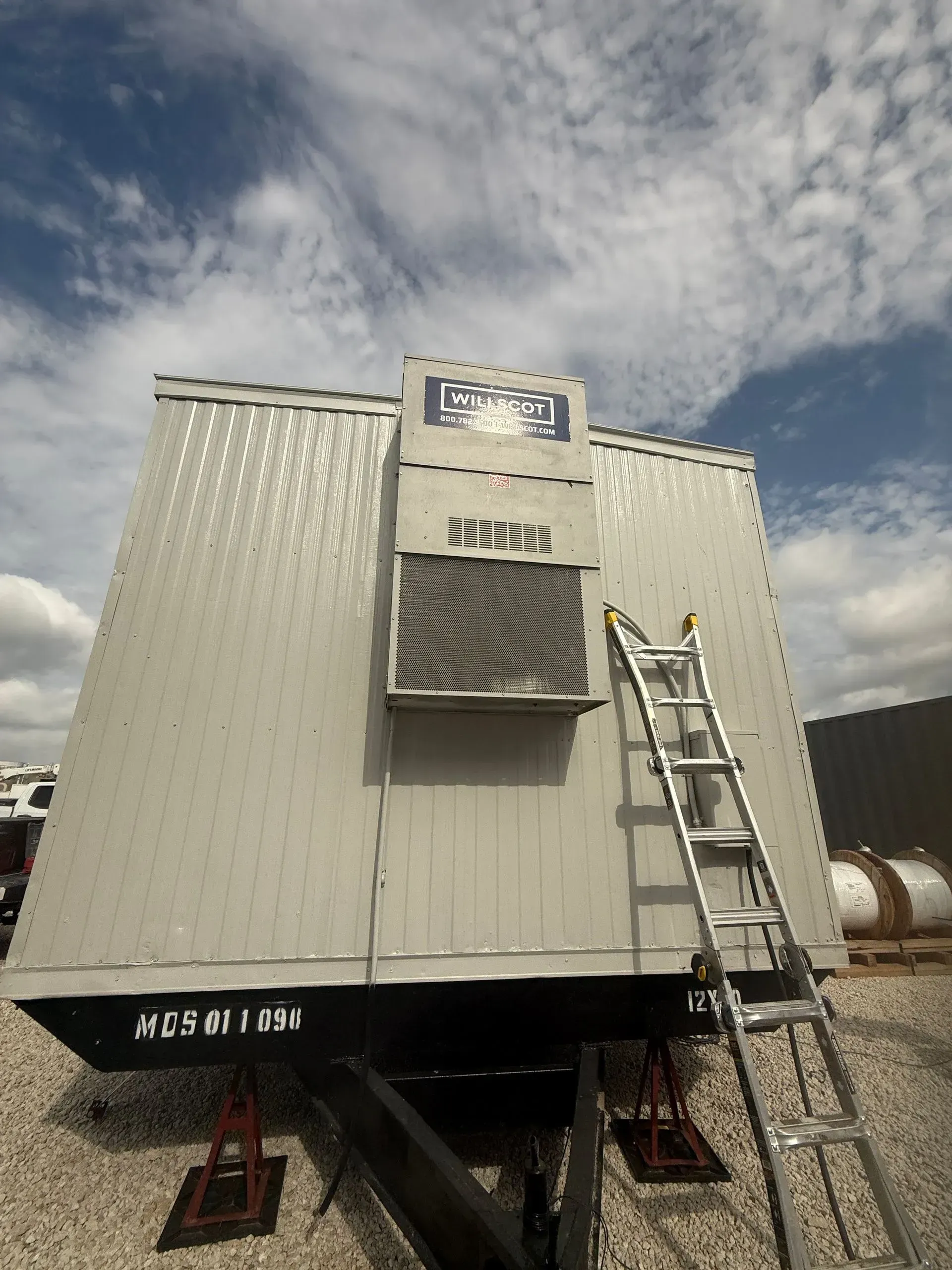 White portable building on stilts with ladder and vent against a cloudy sky