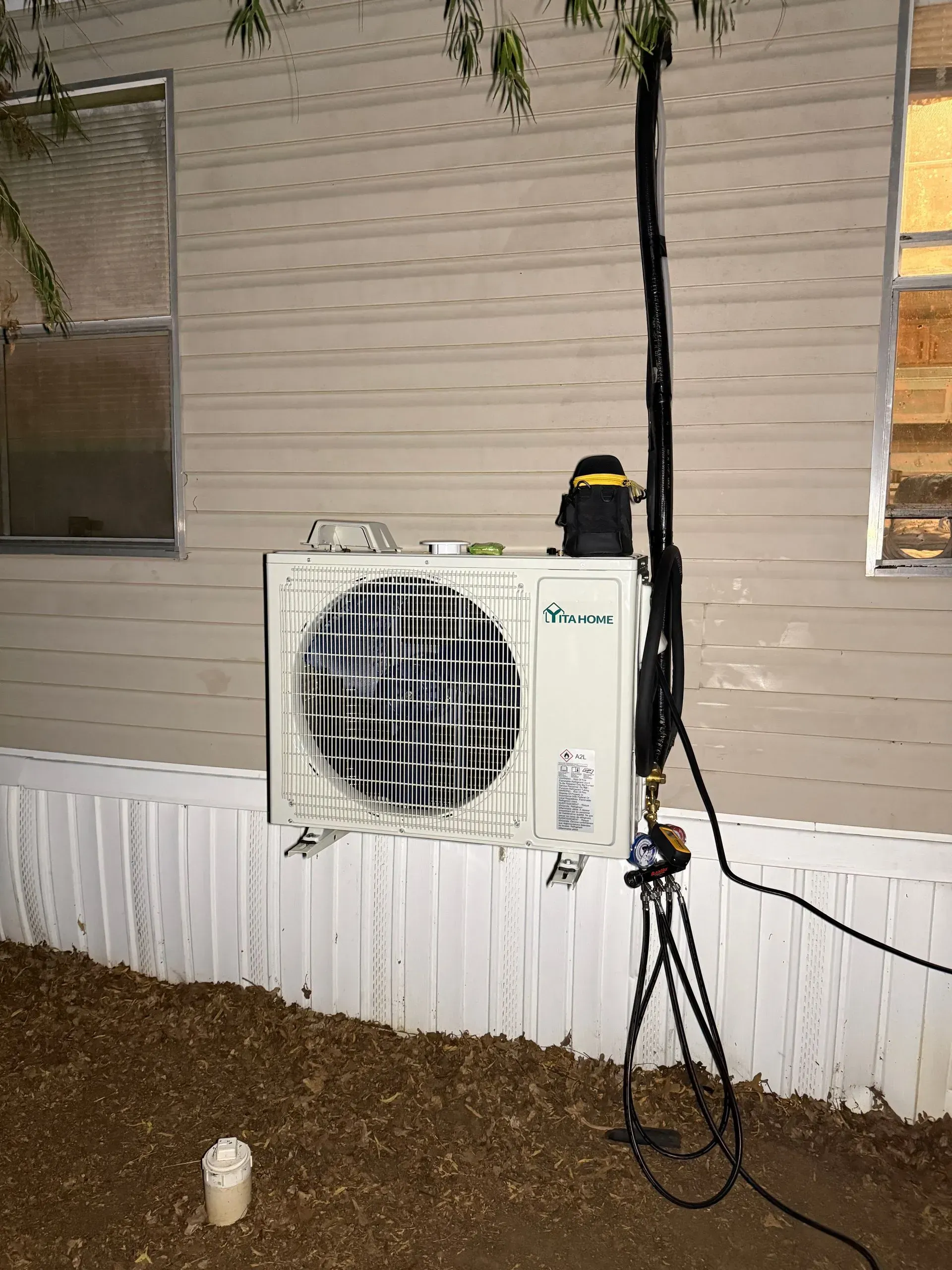 Outdoor wall-mounted HVAC unit beside a house, with a tripod light stand and extension cord nearby.