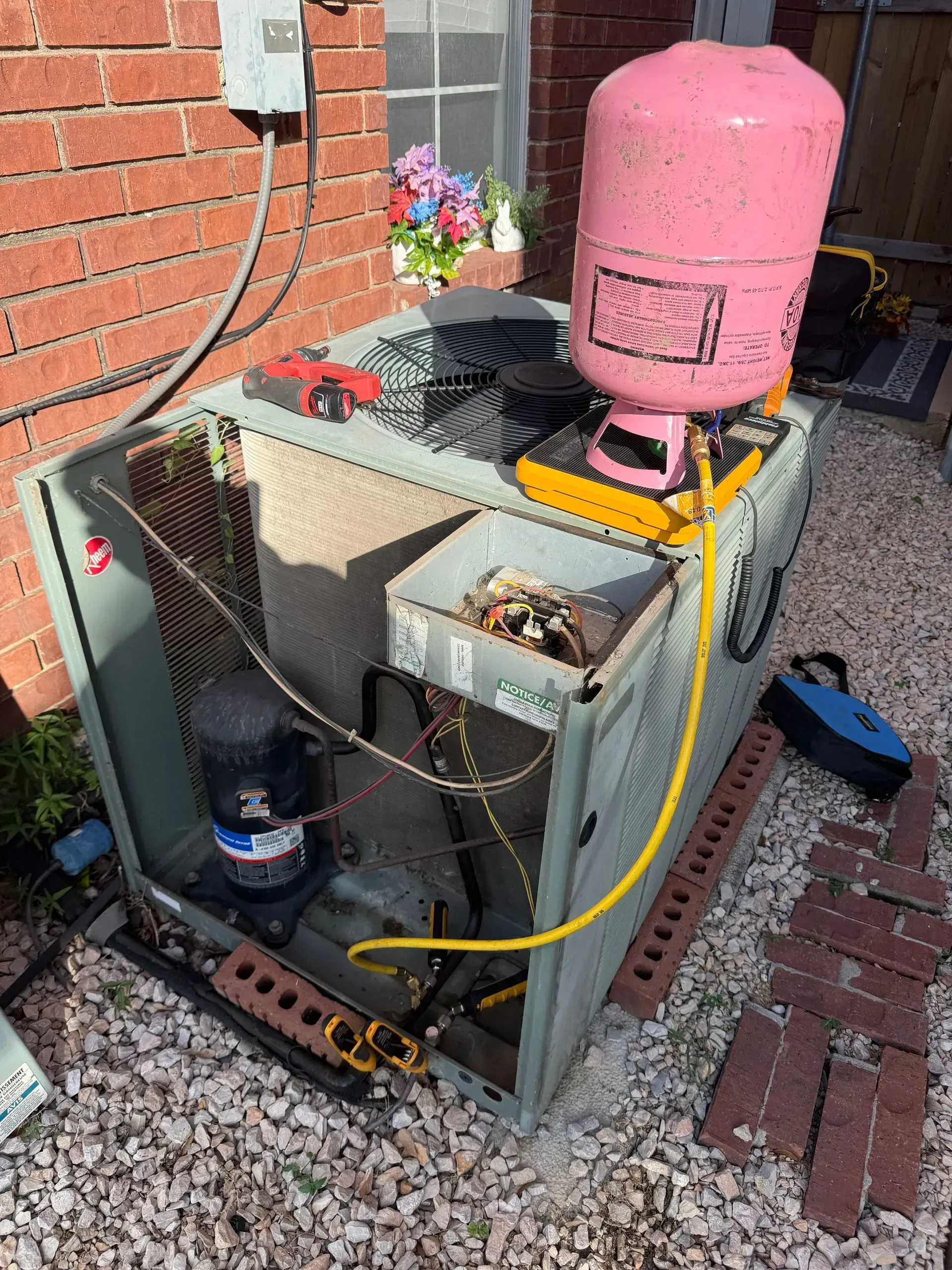Outdoor HVAC unit beside brick wall with pink tank on top and yellow hose attached, on brick pavers.