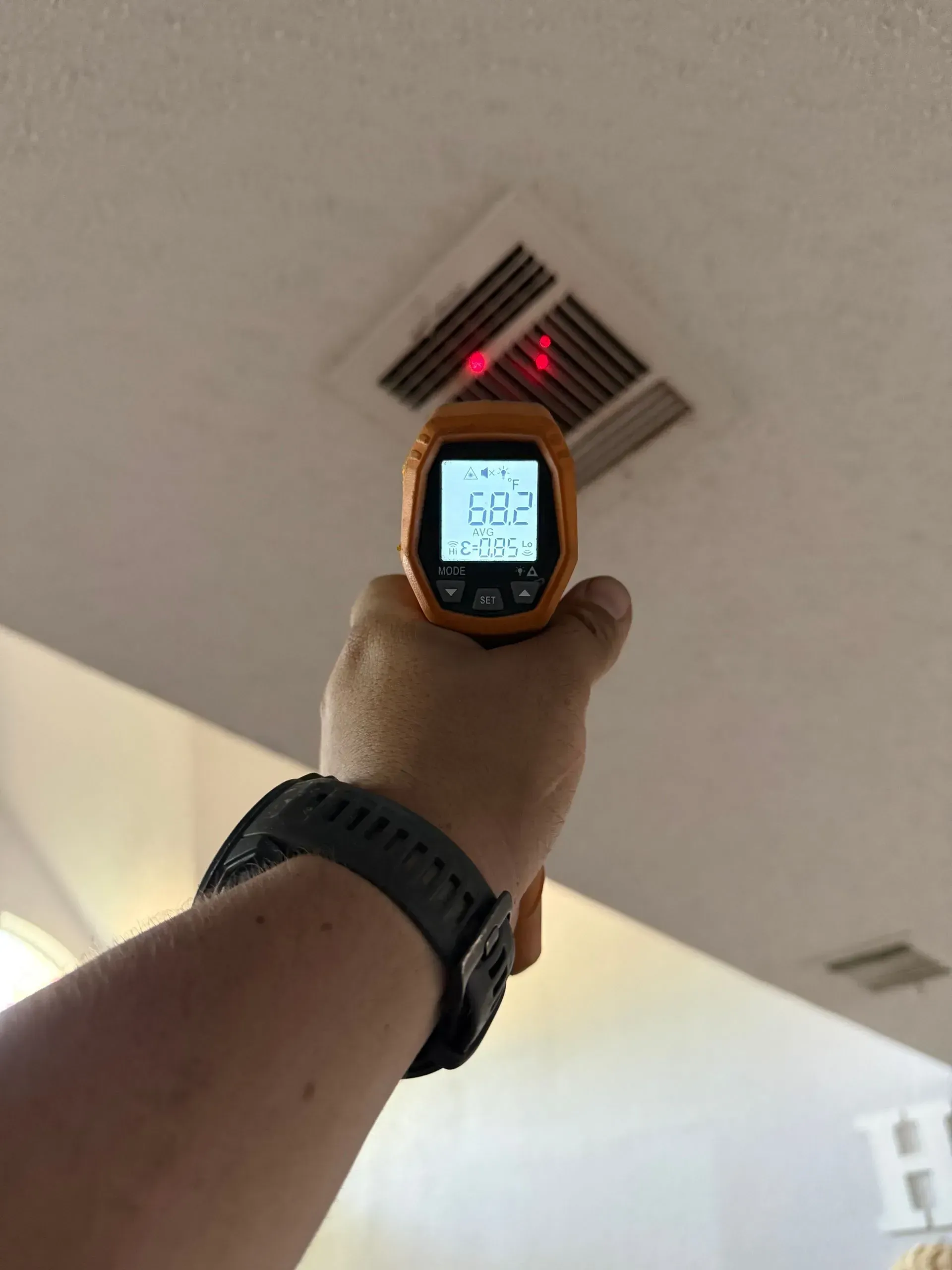 Hand holding a digital thermometer up toward a ceiling vent with red airflow arrows and a gray ceiling.