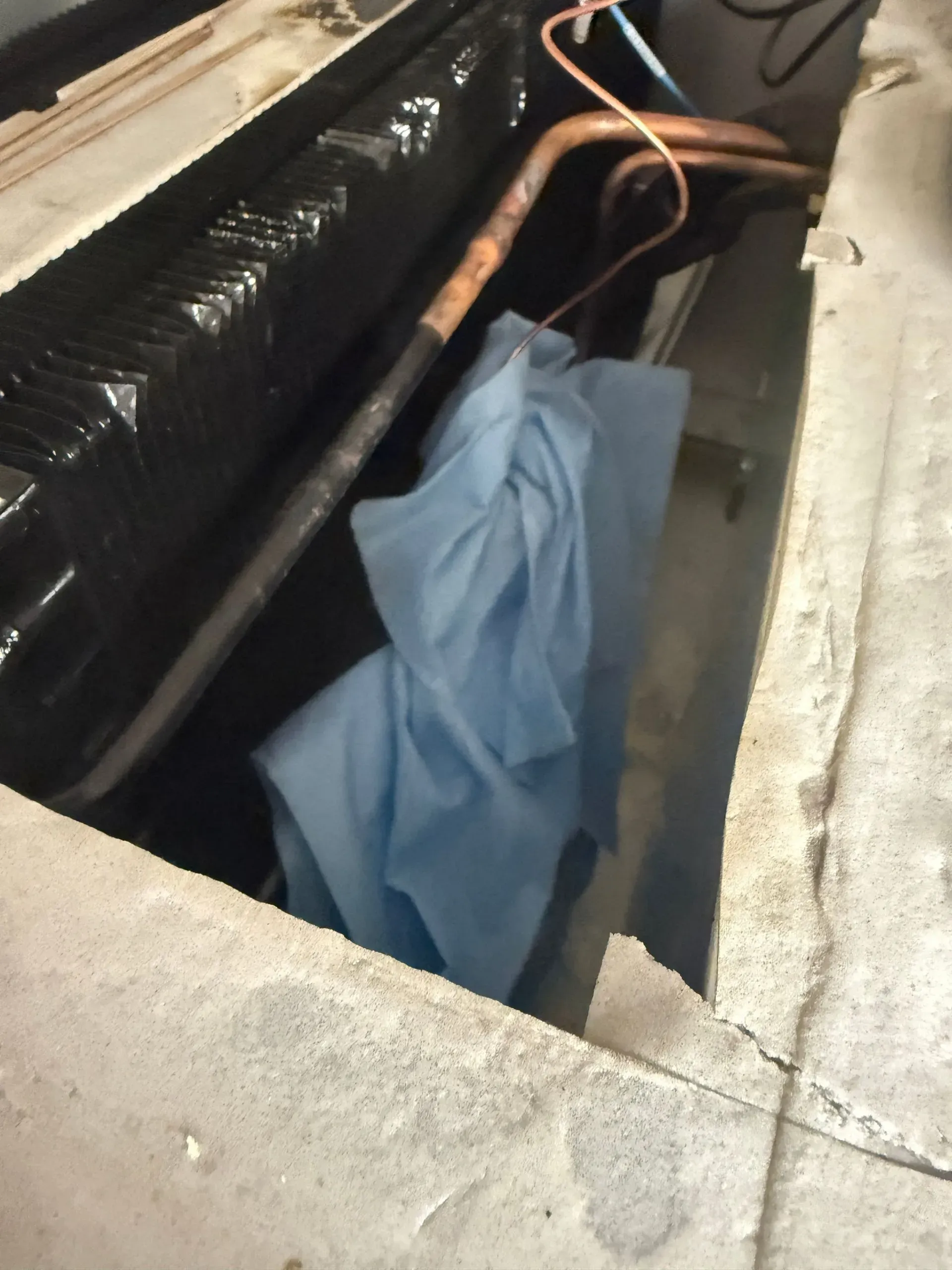 Open floor hatch showing black pipes, orange wires, and a blue cloth draped inside a narrow concrete cavity