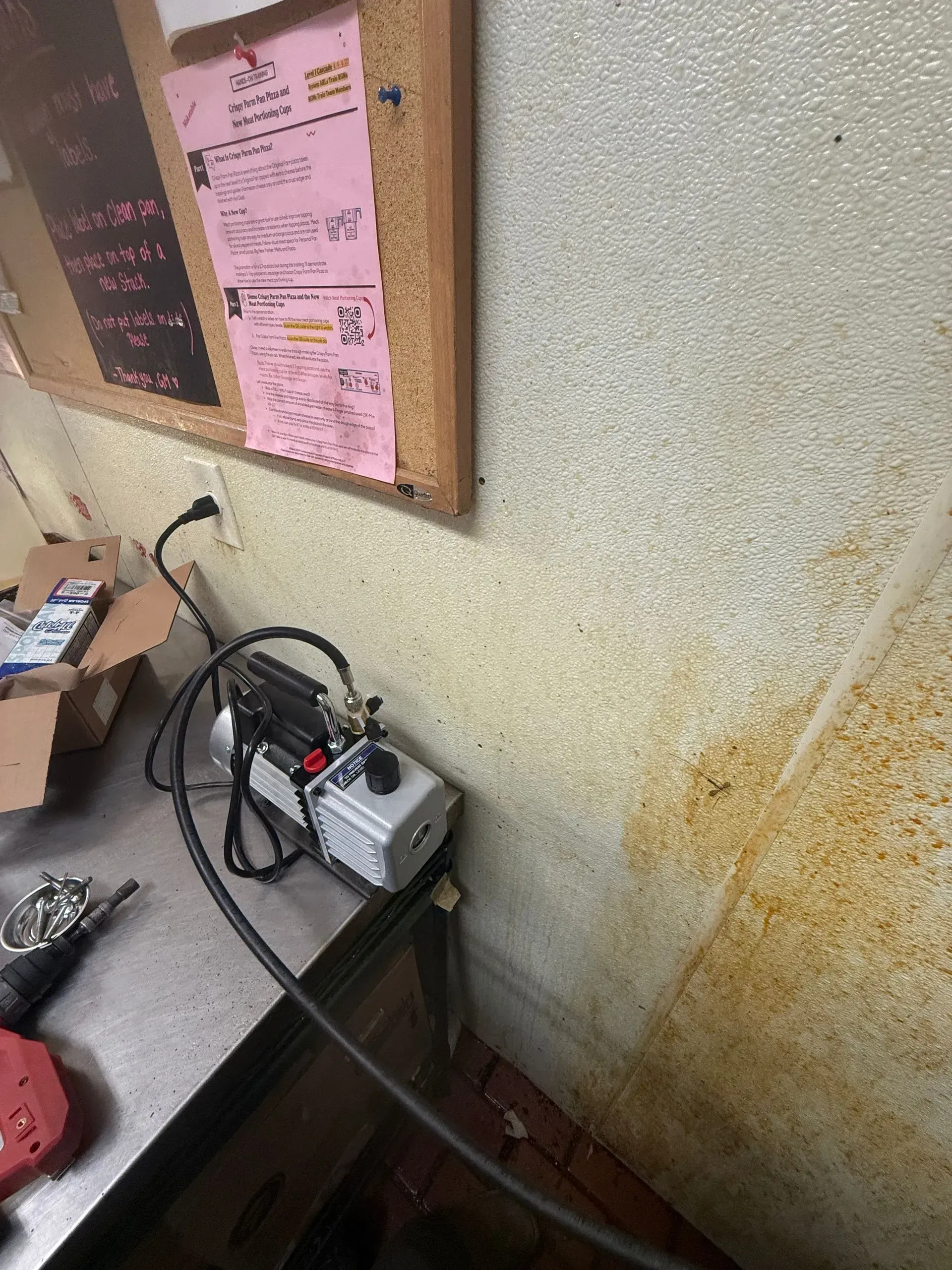 Cluttered desk with a small machine, cables, and a bulletin board against a textured wall.
