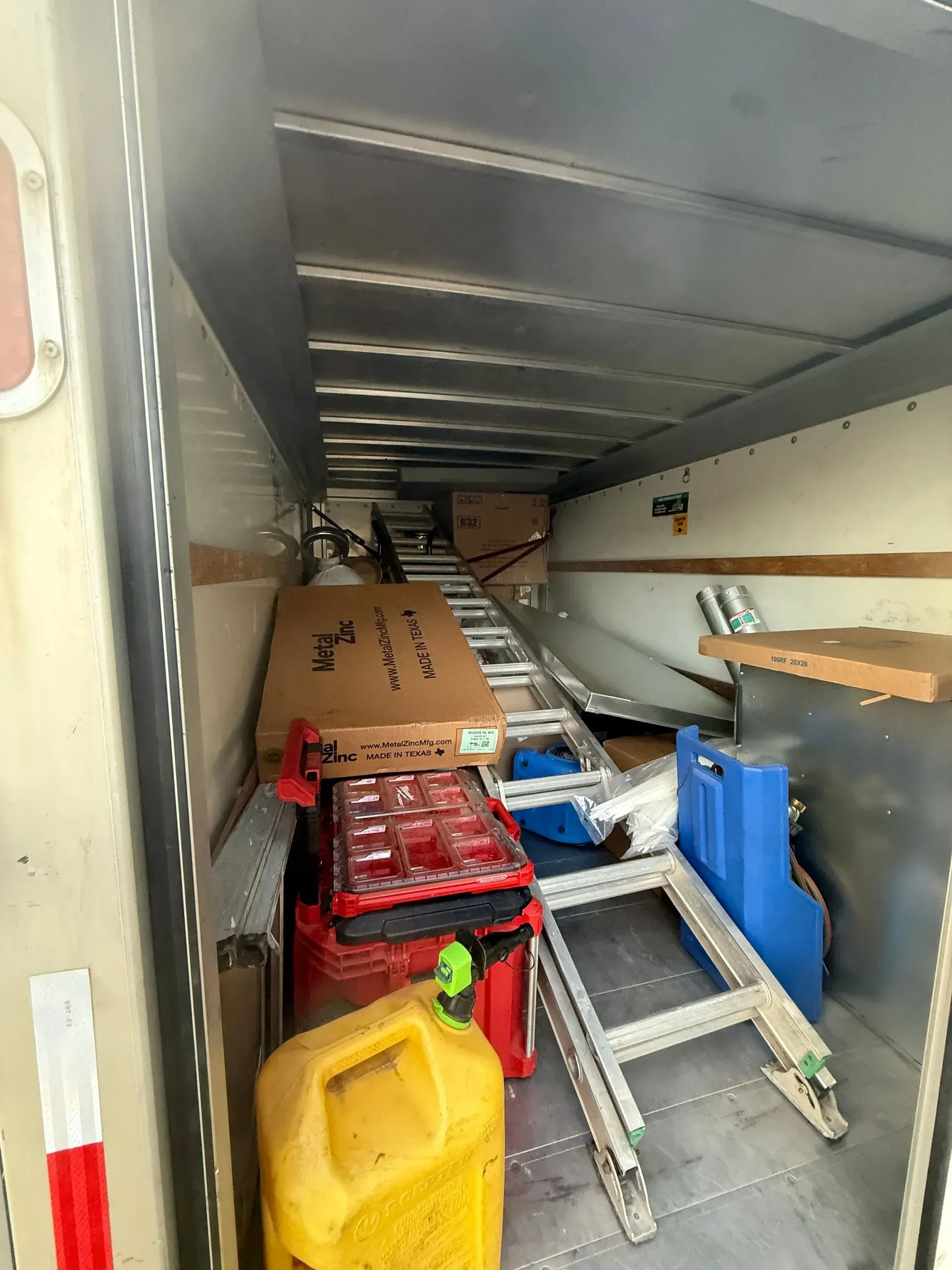 Cluttered truck trailer interior with boxes, crates, tools, a ladder, and a yellow bucket stacked along the floor