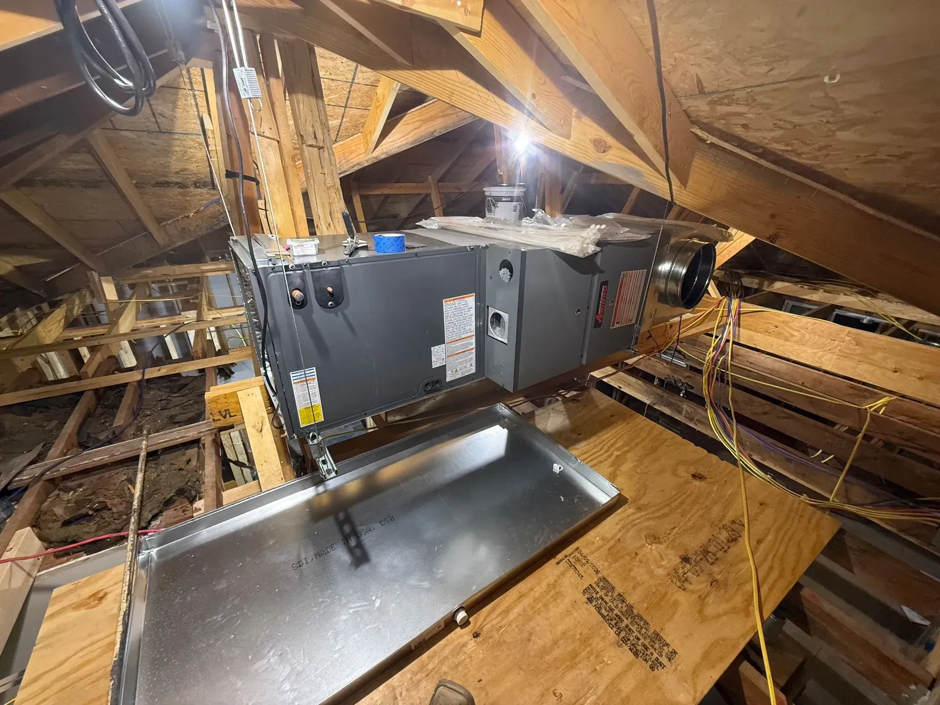 Attic HVAC unit and metal ductwork among exposed wooden rafters with insulation.