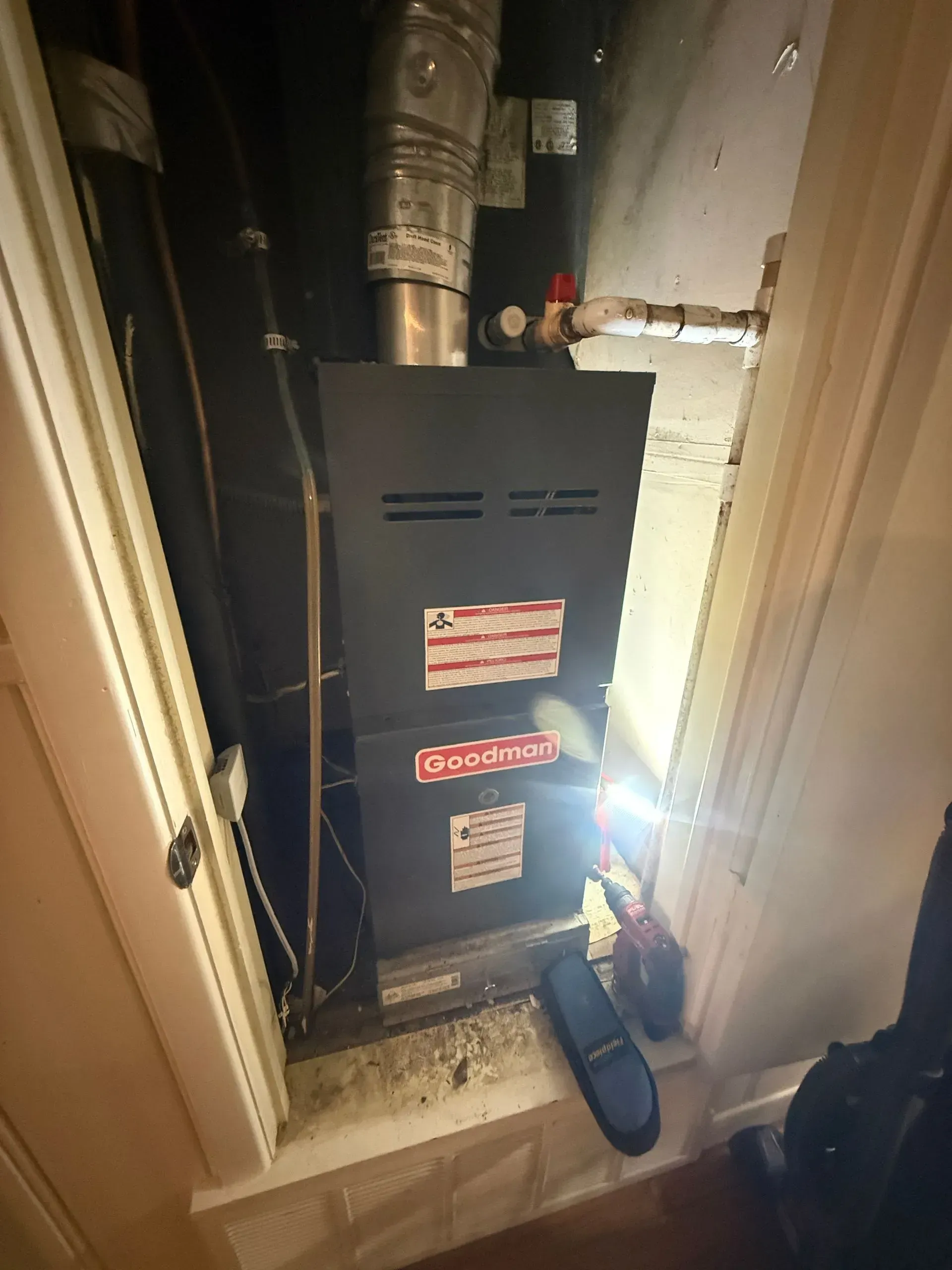 Black furnace in a narrow utility closet with exposed ducts and a flashlight on the floor