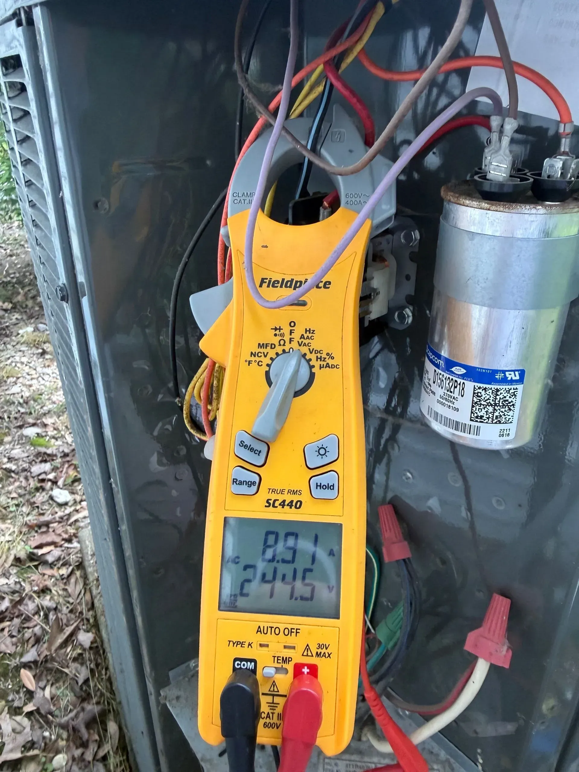Yellow digital multimeter clipped inside an electrical panel, with red and black leads connected to a capacitor.