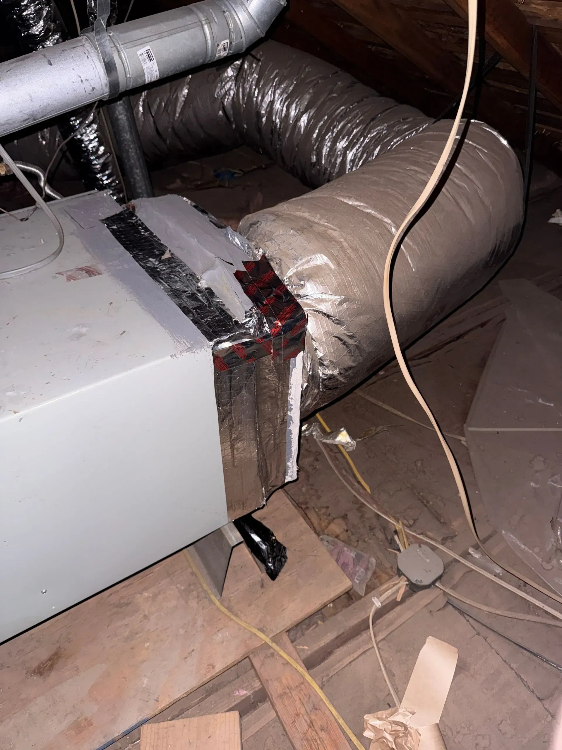 Damaged HVAC duct connected to a metal unit, with torn insulation and debris on a dusty floor.