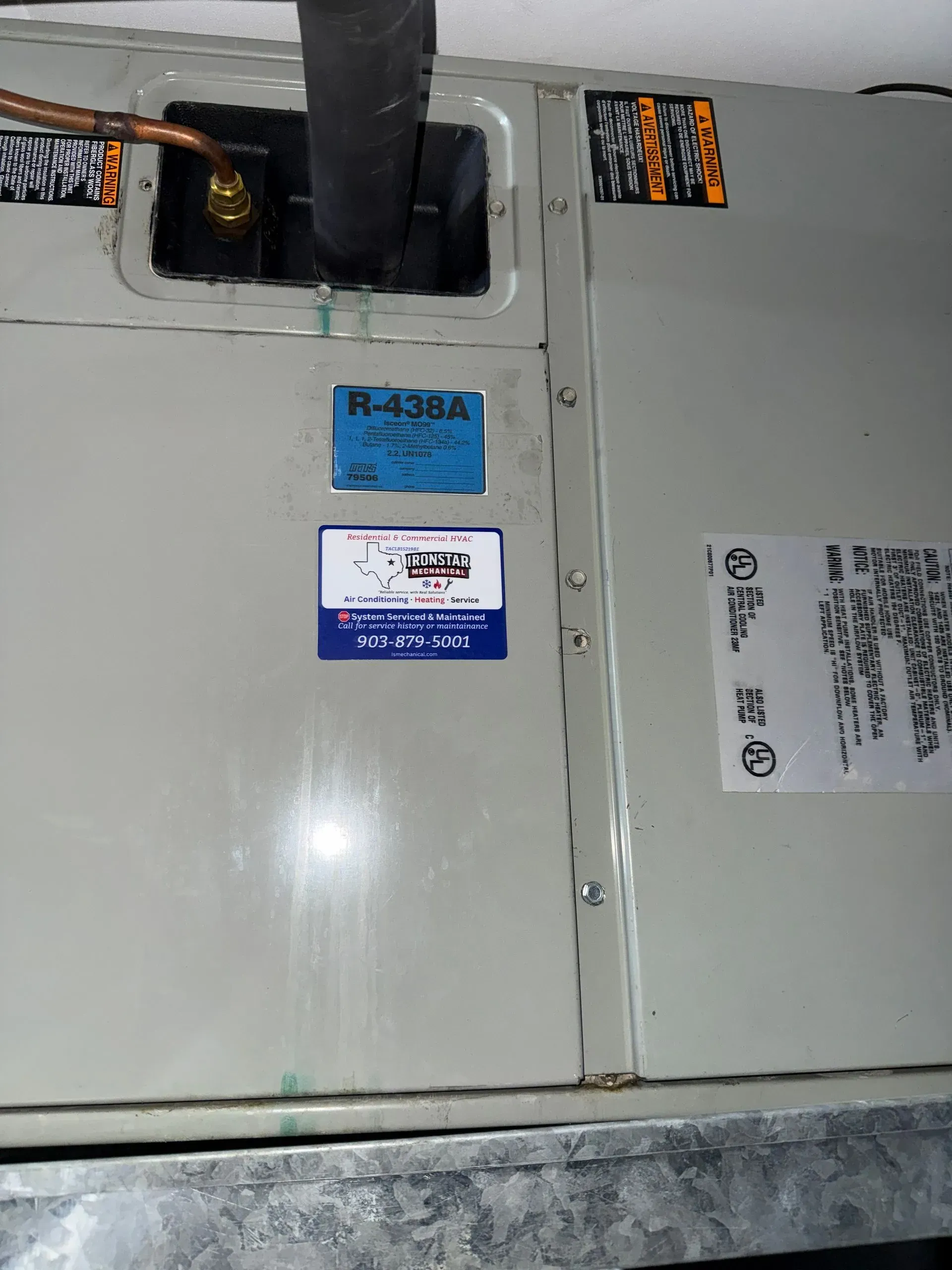 Gray electrical cabinet with labels and black piping/ducts on top, mounted against a wall.