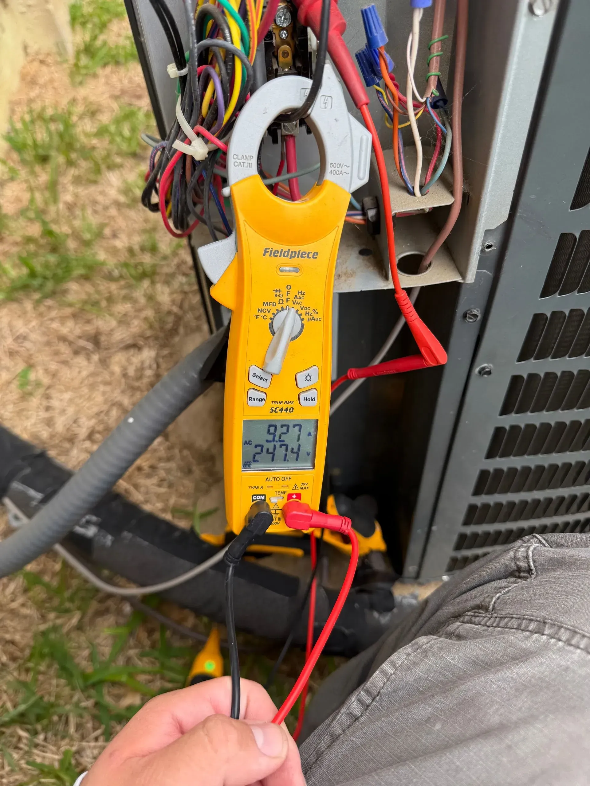 Yellow clamp meter measuring wires inside an outdoor HVAC unit while a hand holds red and black probes