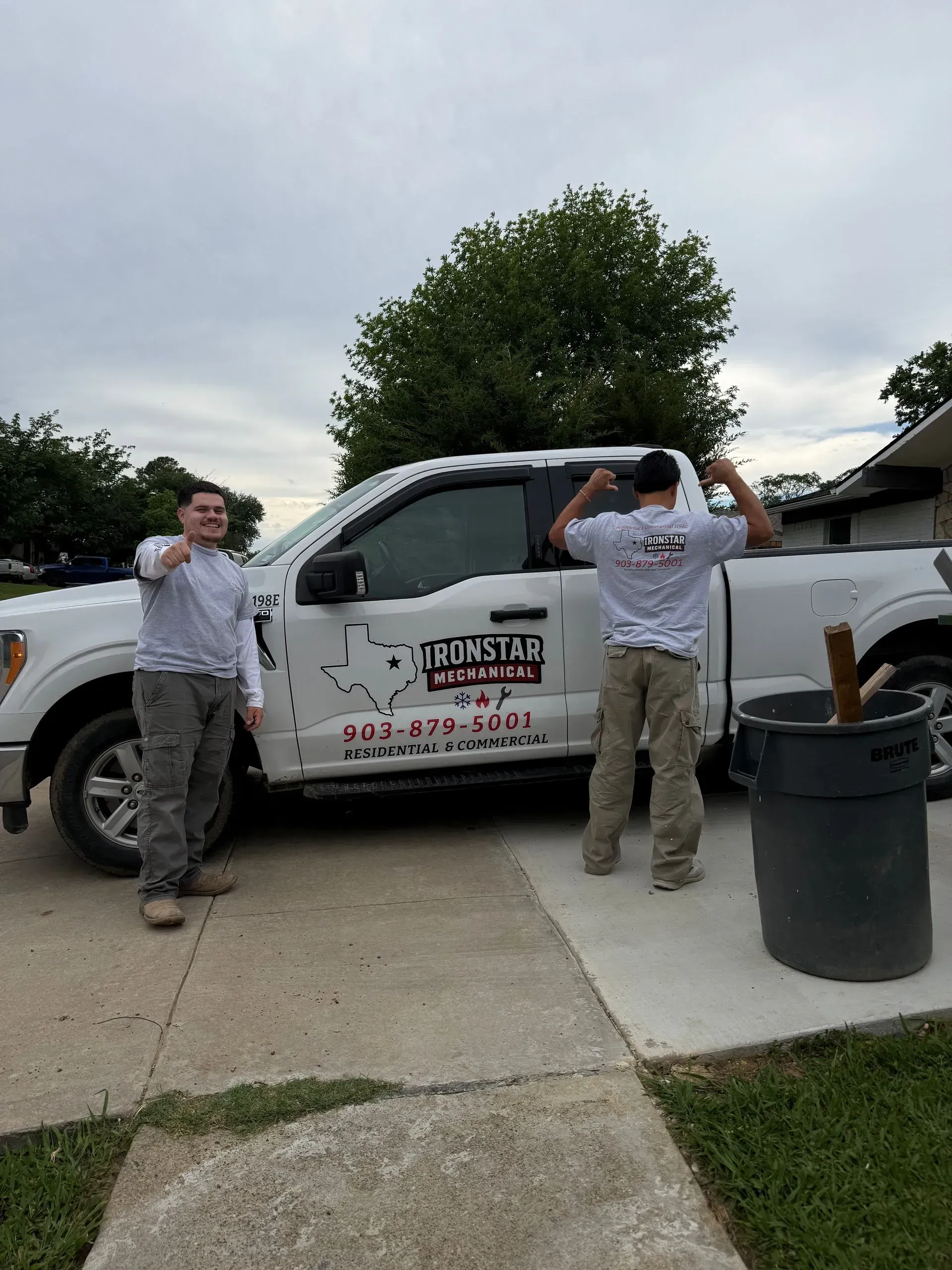 Two people standing beside a white utility truck in a driveway, one posing with hands on hips, one shielding eyes.