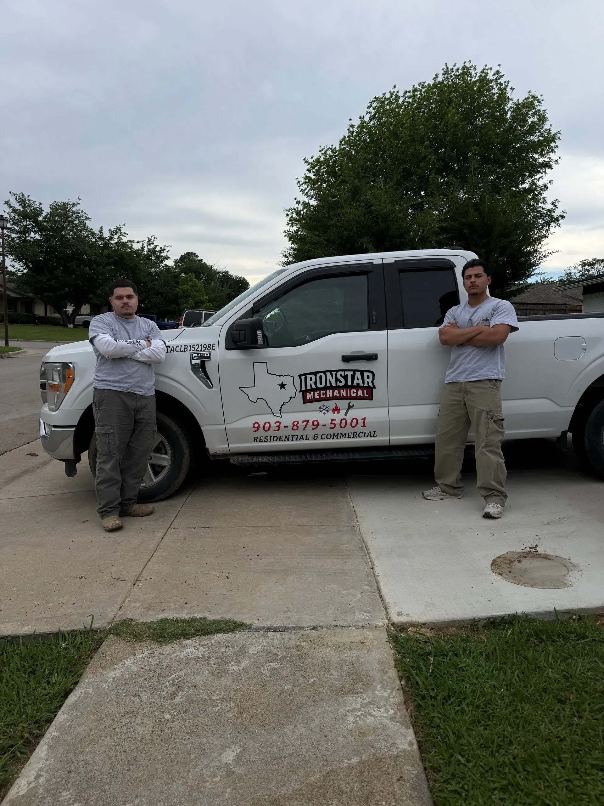 Two workers leaning against a white service truck parked in a driveway with a tree behind it.