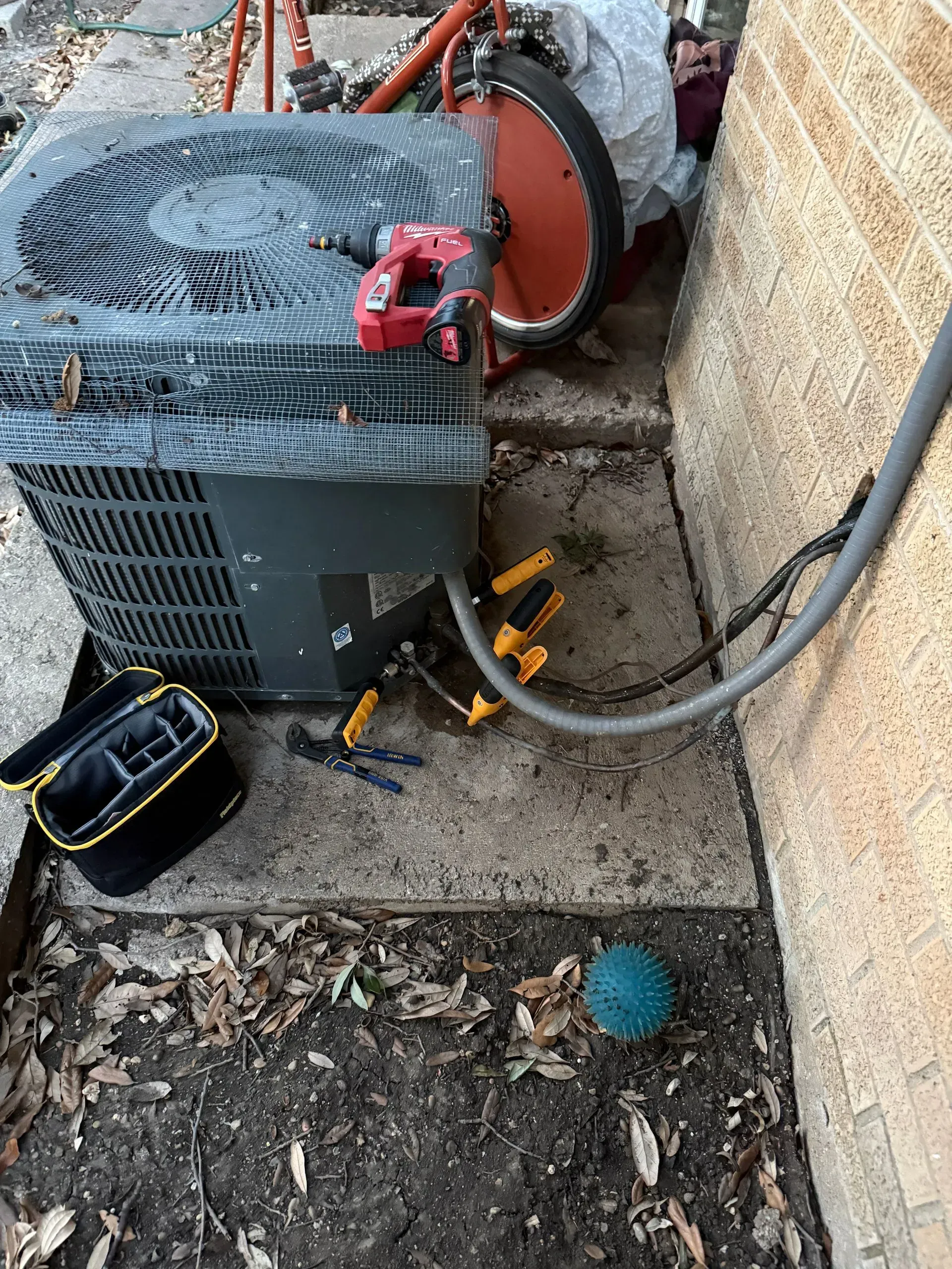 Outdoor HVAC unit beside a brick wall, with hoses, tools, and debris on the ground.