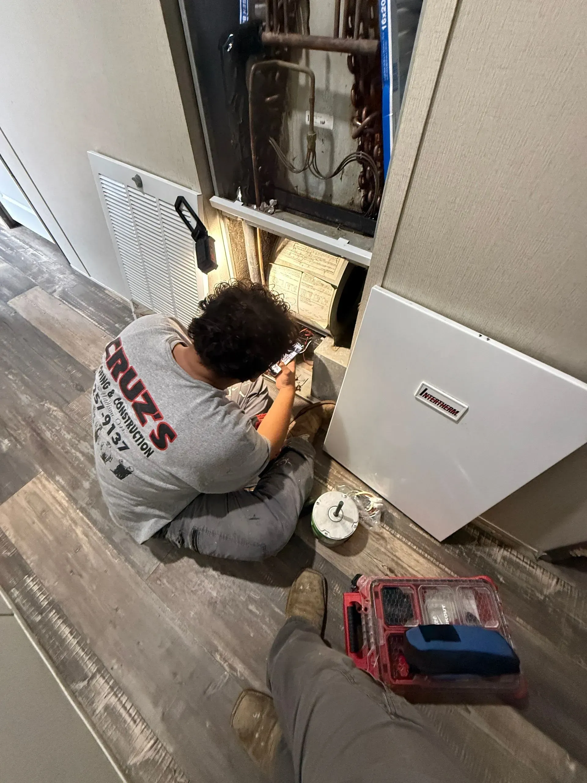Technician kneels by an open electrical panel, working with tools in a home hallway.