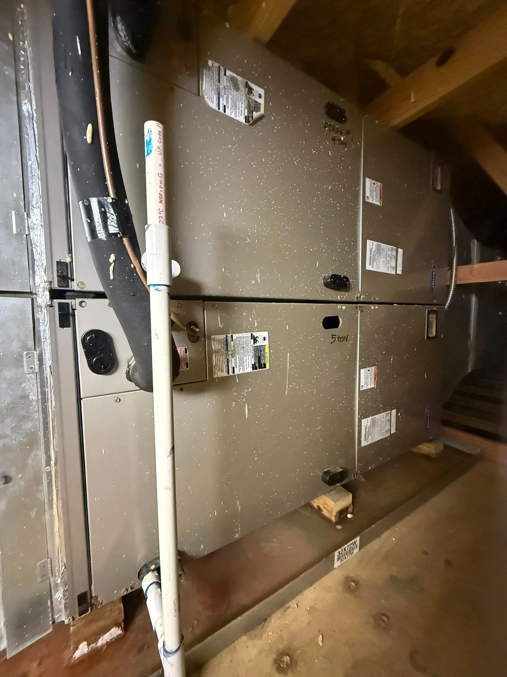 Large metal HVAC units in a narrow mechanical space with exposed pipes and a concrete floor.