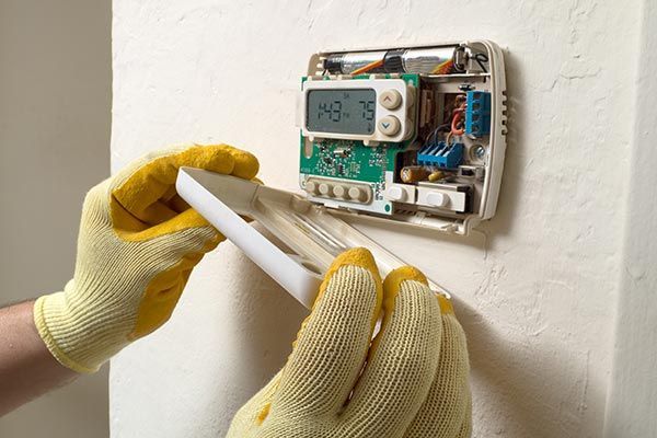 Gloved hands installing a white thermostat on a wall with exposed wiring