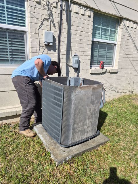 Worker servicing an outdoor HVAC unit beside a house window, leaning over the open top panel.
