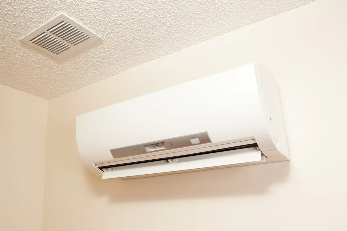 Wall-mounted white air conditioner near a ceiling vent in a beige room.