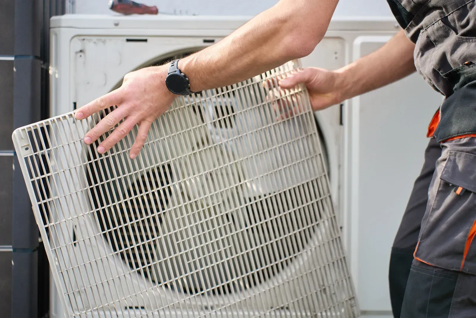Two people lifting a large white appliance filter panel beside an open unit outdoors