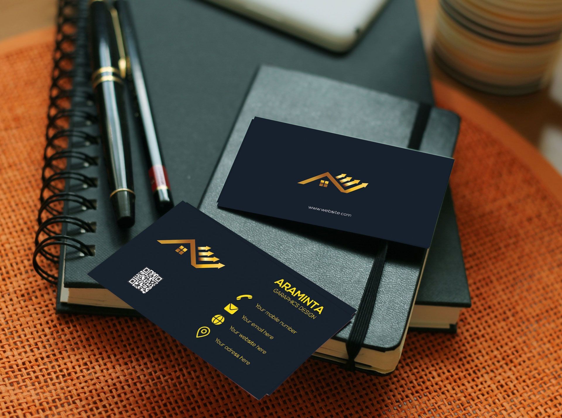 Two dark blue business cards with gold logos and text placed on top of a black notebook with pens on a textured surface.