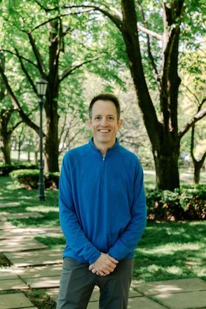 Person standing on a tree-lined sidewalk in a blue jacket, smiling at the camera.