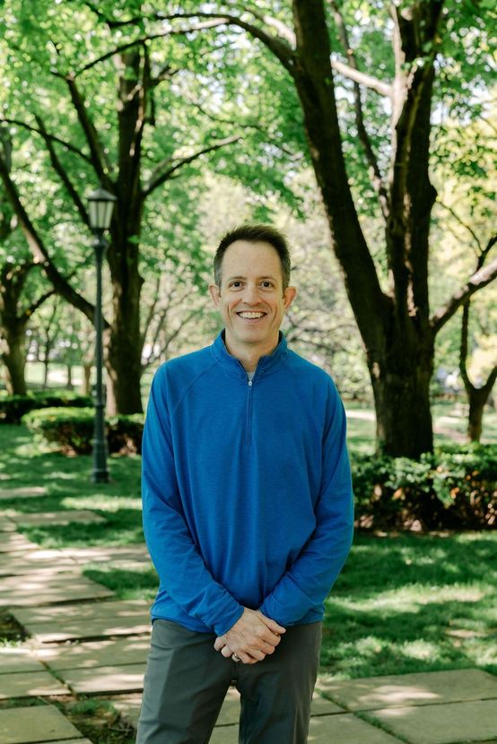 Man standing on a tree-lined path in a park, wearing a blue jacket and smiling.