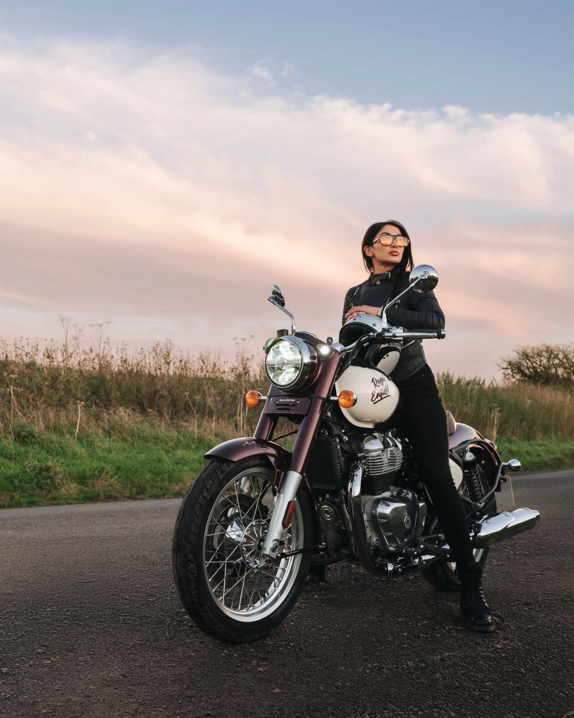 Pessoa sentada em uma motocicleta preta em uma estrada rural ao pôr do sol, com campos e nuvens rosadas ao fundo.