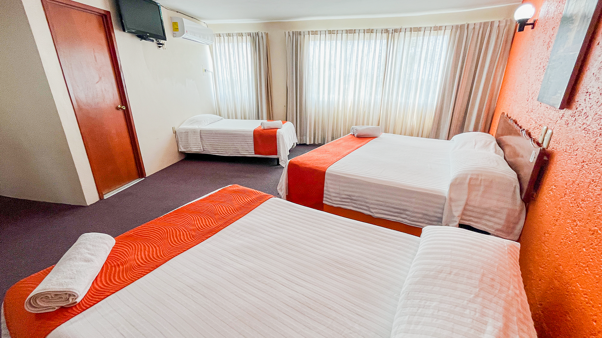 A hotel room with two large beds in the foreground and a smaller single bed in the back near a window with curtains.