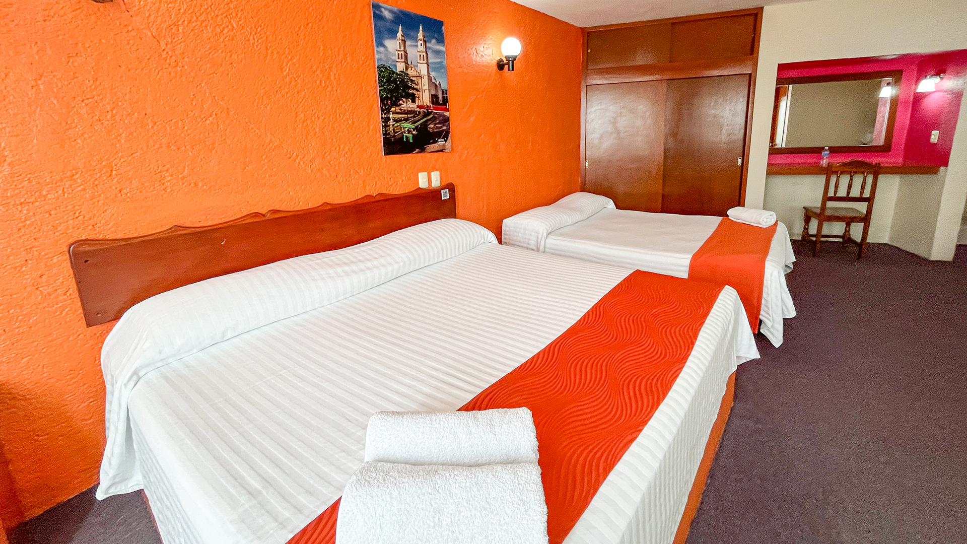 A hotel room with two beds, orange walls, purple carpet, and a wooden desk with a mirror.