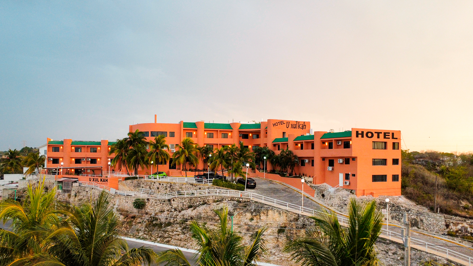 A three-story orange hotel with a green roof sits on a rocky, tree-lined hill under a sunset sky.