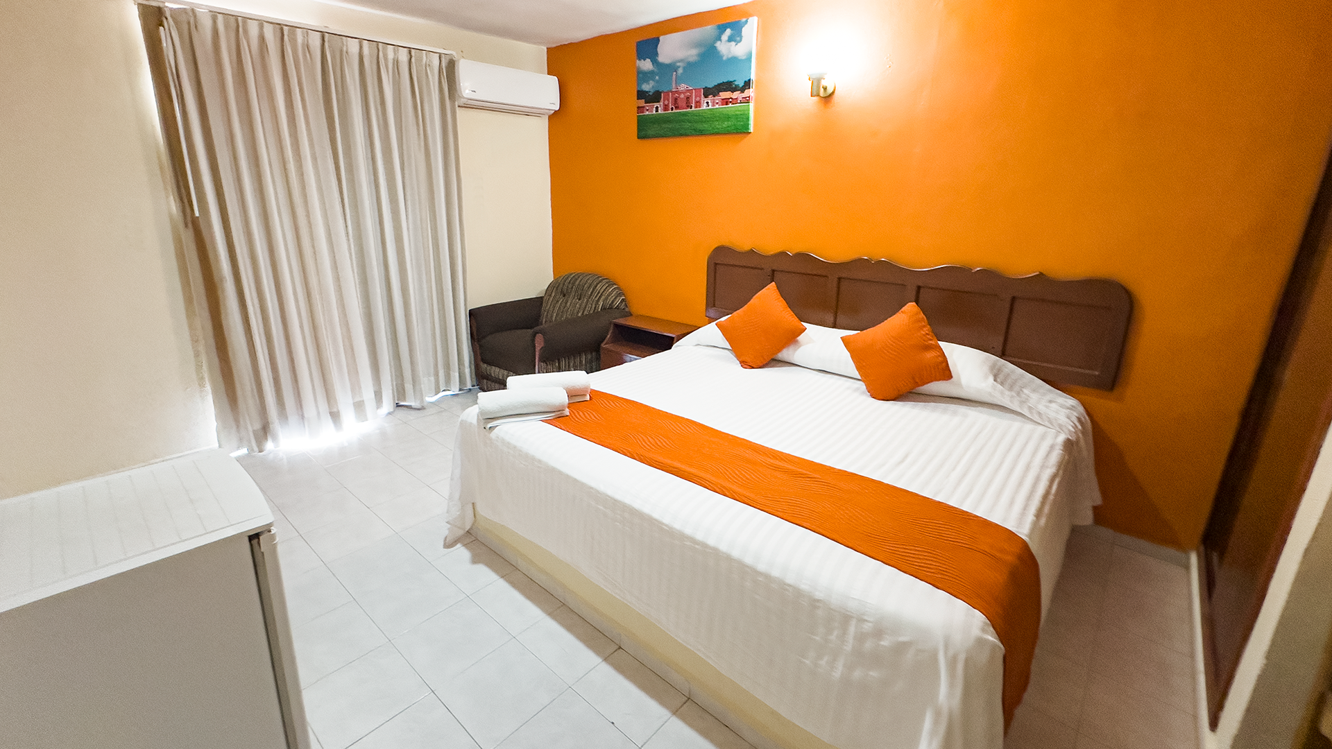A hotel room with a queen bed featuring white linens and an orange runner, set against an orange accent wall.
