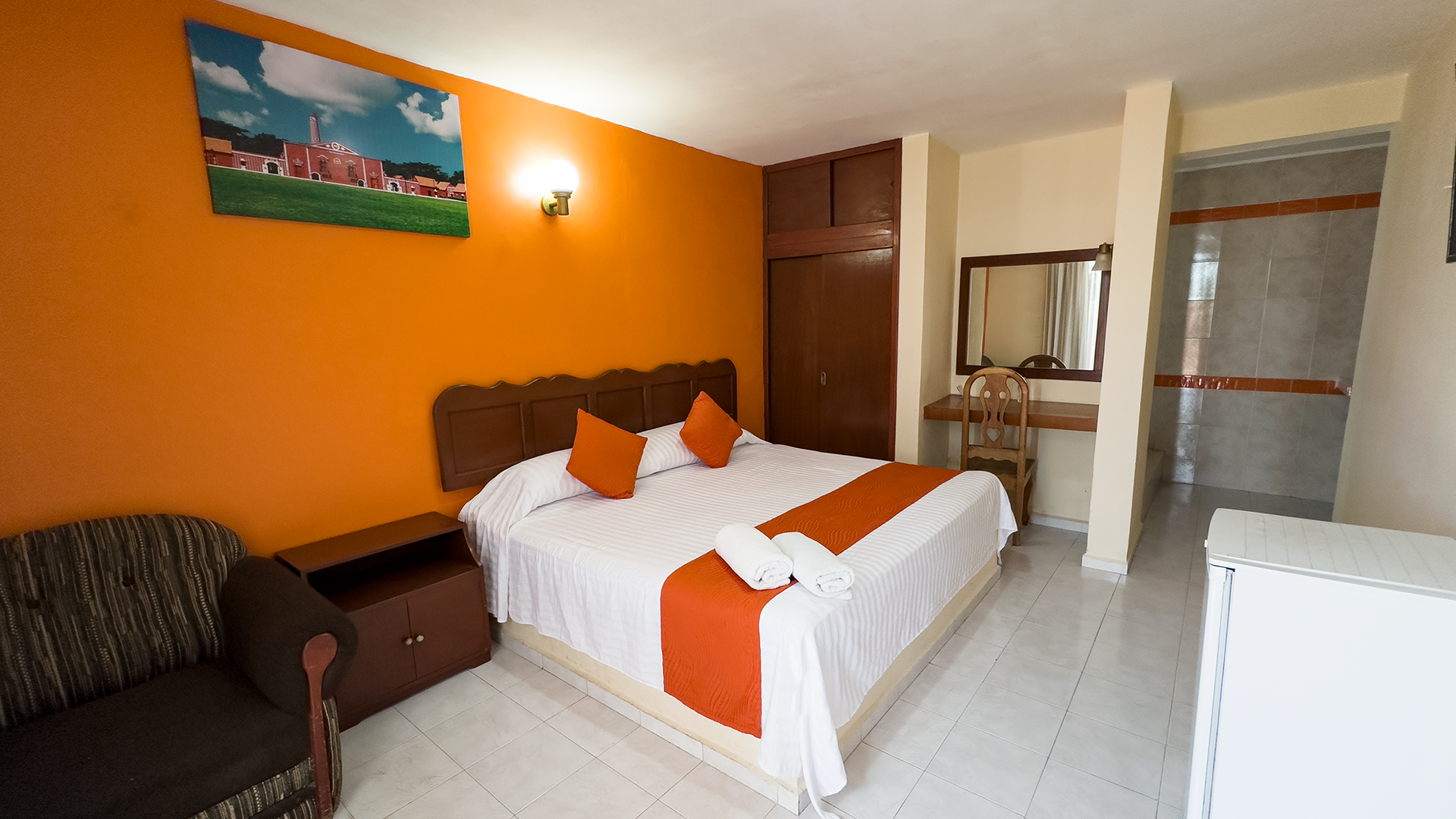 A bedroom with a bed featuring orange accents, an orange wall with a picture, a wooden cabinet, and a chair by a vanity.