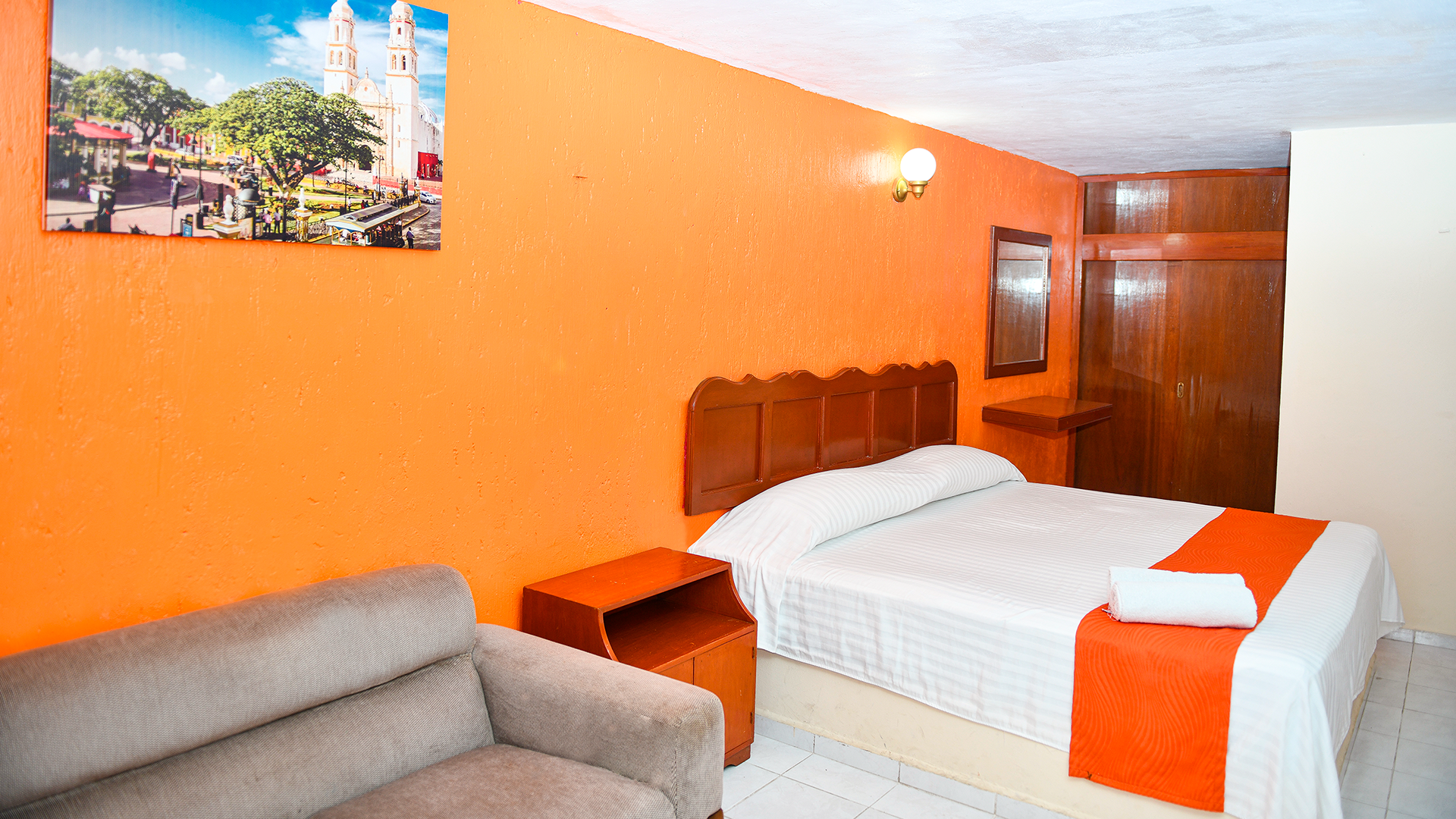 A hotel room with bright orange walls, a double bed with a white and orange runner, a brown sofa, and a wall painting.