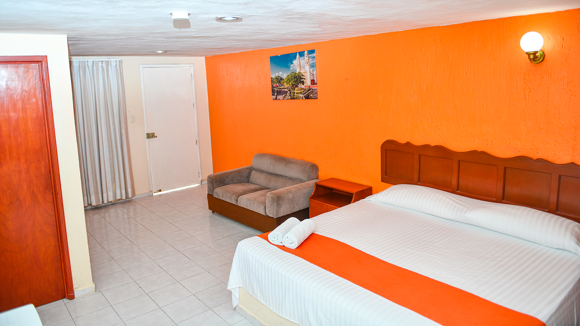 A hotel room with a white bed, an orange accent wall, a small sofa, a wooden headboard, and white tile floors.