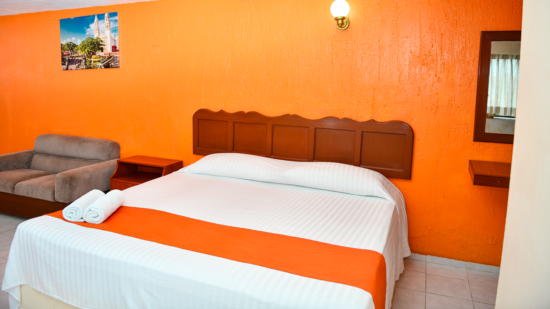 A hotel room with a bed featuring white linens and an orange runner, a gray sofa, and bright orange walls.