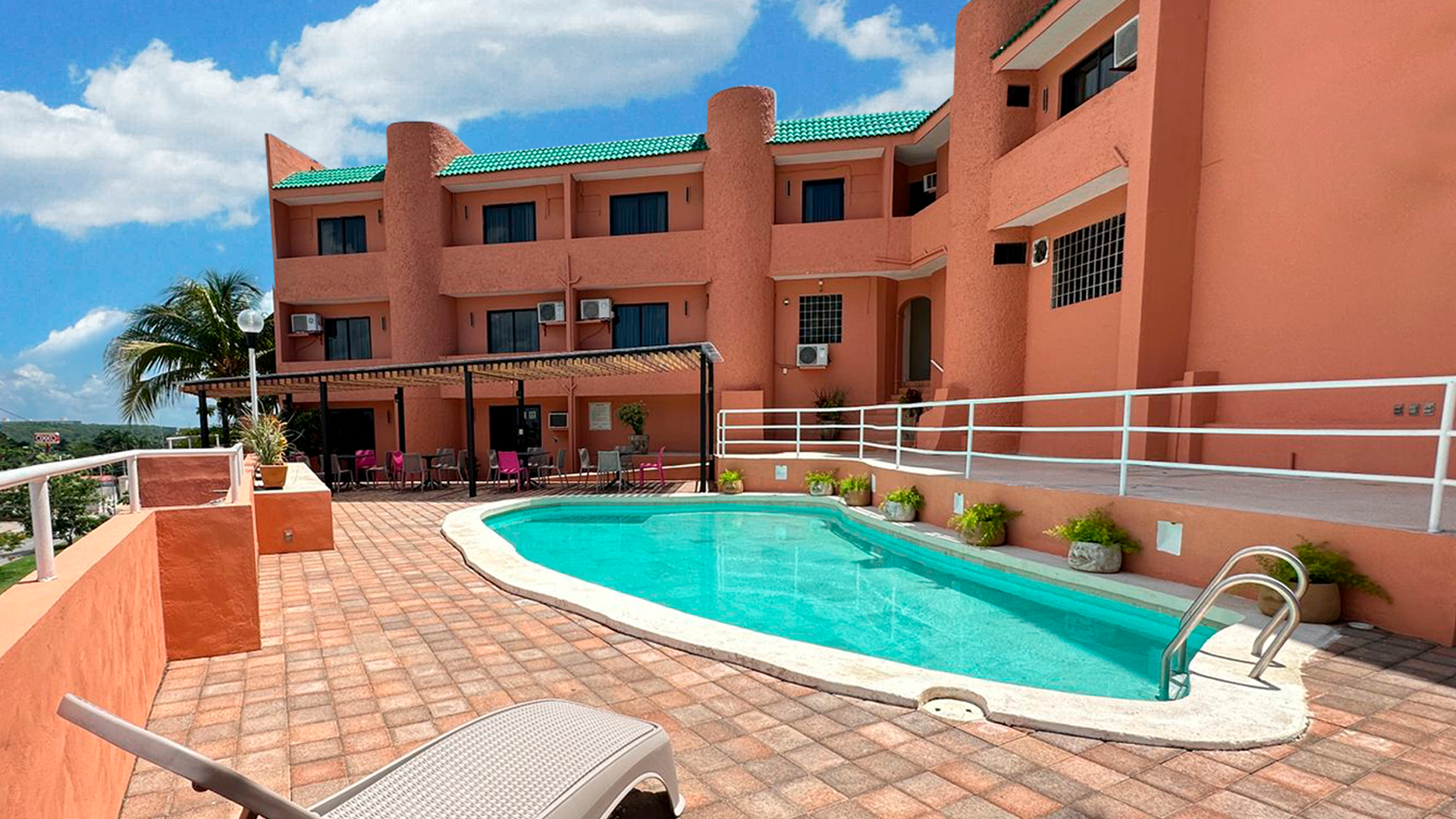 A swimming pool sits on a brick patio in front of a multi-story, salmon-colored building under a bright blue sky.