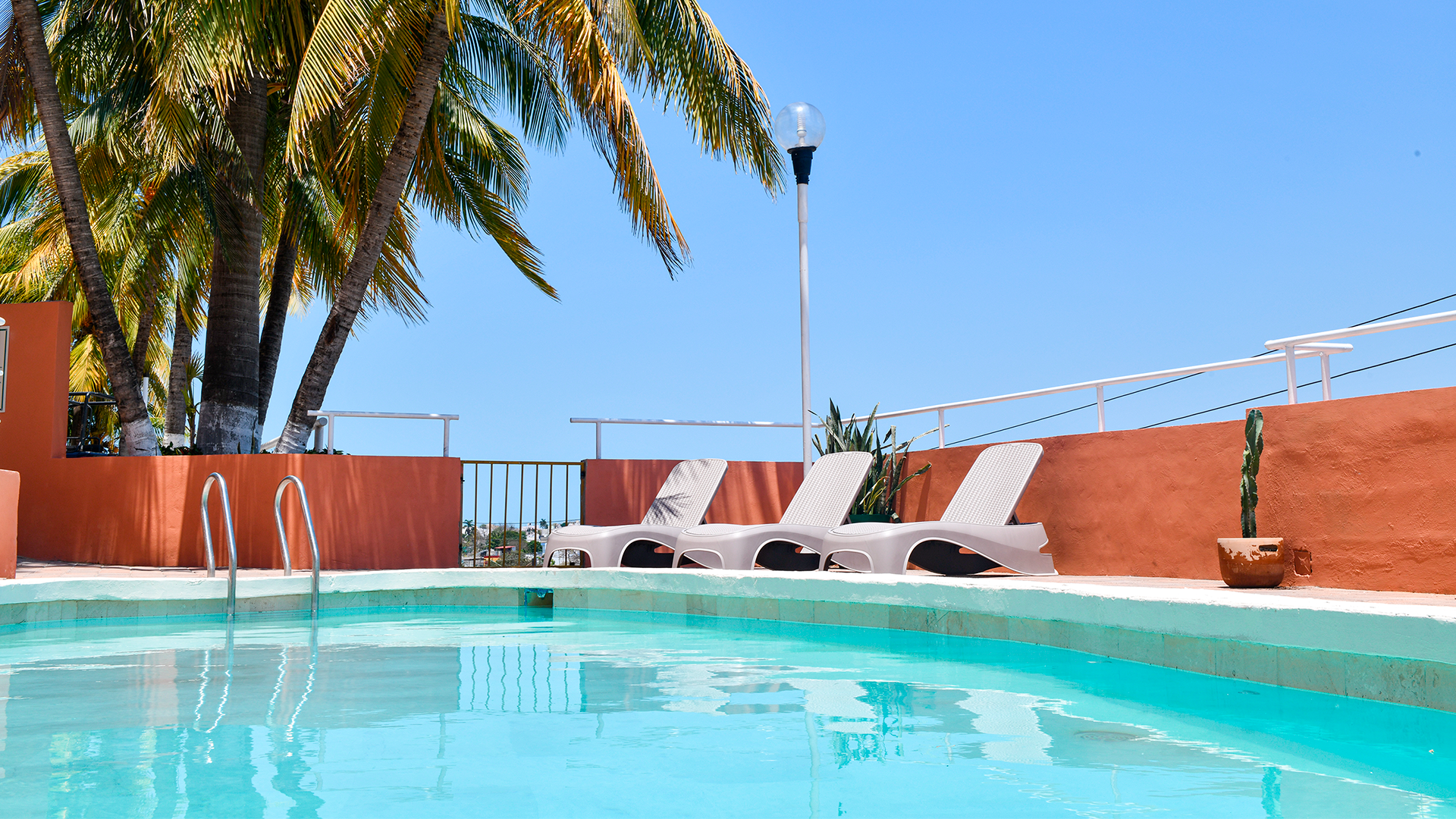 A sunny swimming pool with turquoise water and lounge chairs against an orange wall under palm trees and a clear blue sky.
