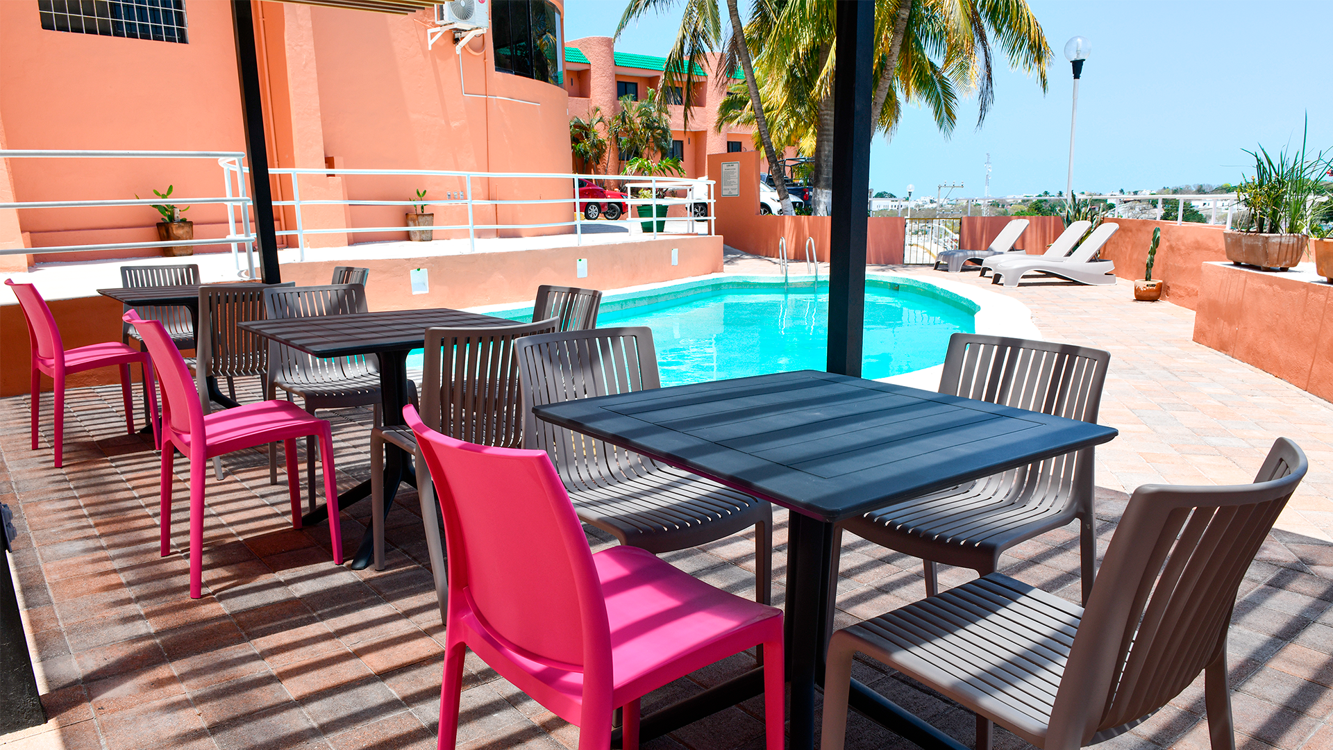 A sunlit patio with tables and chairs near a swimming pool at a resort-style building with peach-colored walls.
